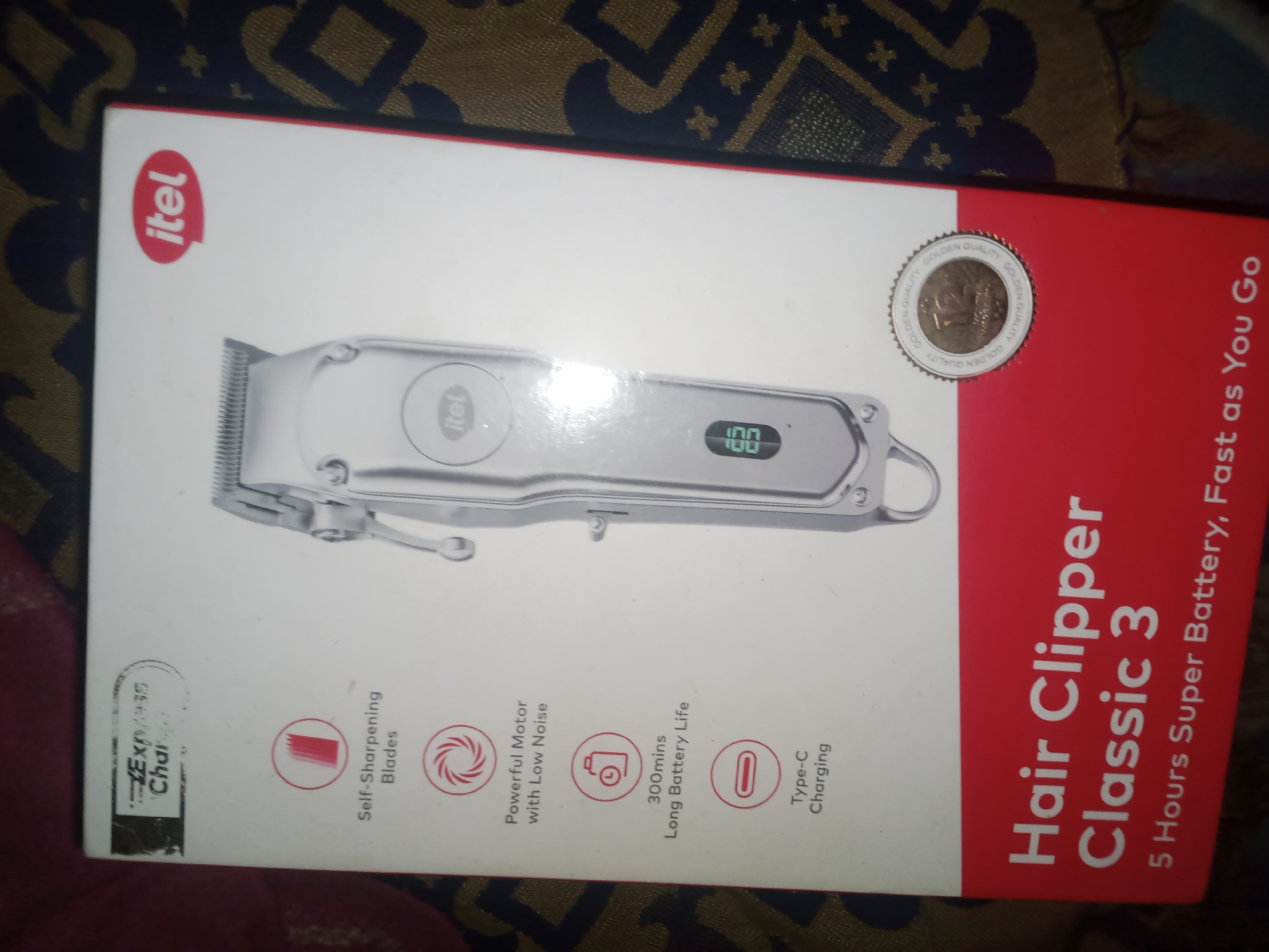 The image displays the packaging of an itel Hair Clipper Classic 3. The box is predominantly white with a red accent on the right side. The itel logo is visible in red on the upper left corner. A depiction of the hair clipper itself is centered on the white portion of the box, showing a silver device with a digital display indicating "100". Below the clipper, icons and text highlight features such as "Self-Sharpening Blades", "Powerful Motor with Low Noise", "300mins Long Battery Life", and "Type-C Charging". A circular emblem with "GOLDEN QUALITY" is present on the upper right of the box. The red section on the right side of the box prominently features the text "Hair Clipper Classic 3" and "5 Hours Super Battery, Fast as You Go". The background of the image appears to be a patterned fabric, possibly a tablecloth or a rug. The image is taken indoors, with the lighting suggesting artificial light sources. There are no people, animals, or specific urban elements of Jauro Yinu, Nigeria visible in this image. The image is a product shot of a hair clipper box.