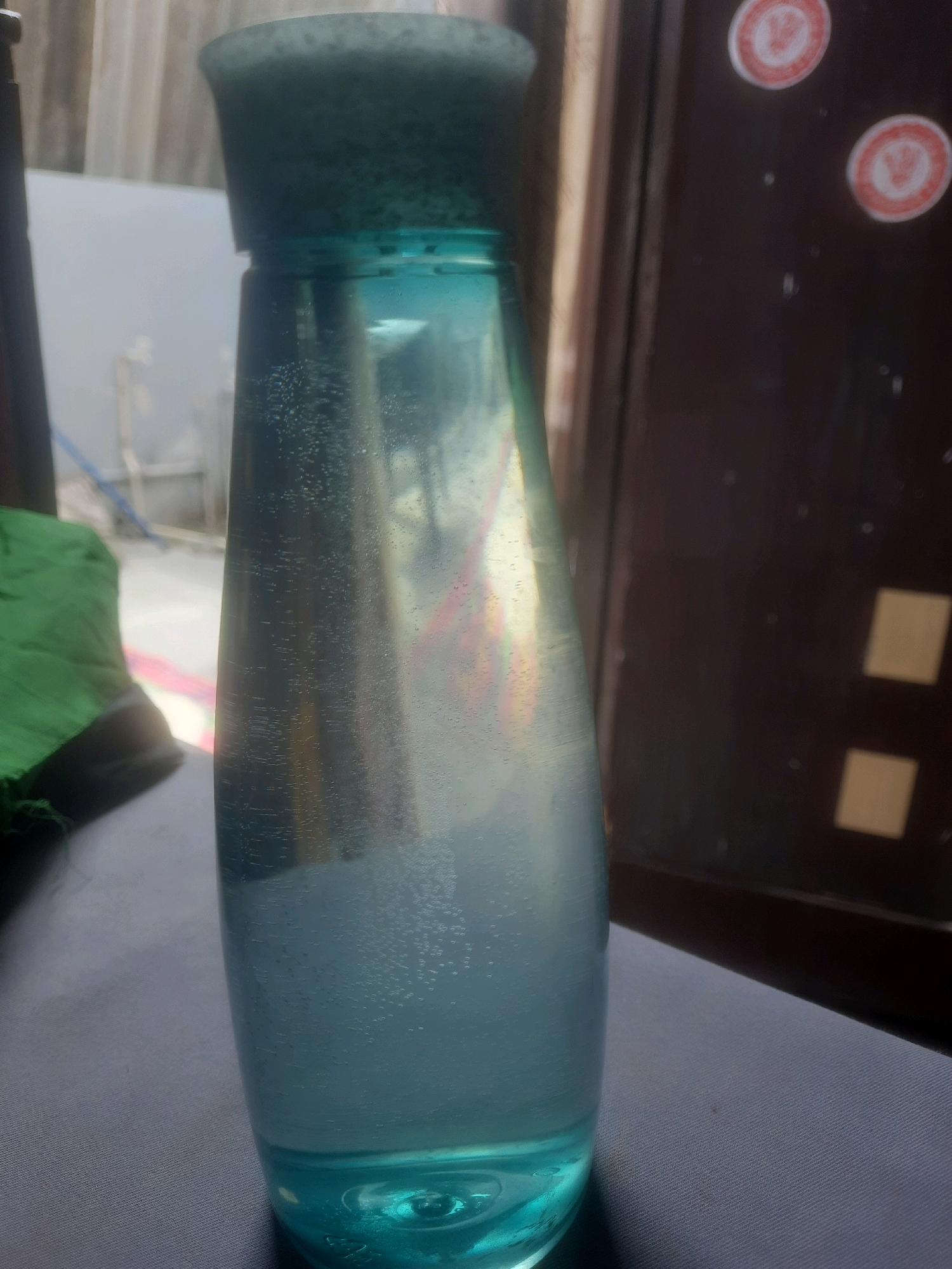 A close-up, vertical, eye-level shot shows a blue-tinted glass bottle with a dark grey cap. The bottle contains a light blue liquid with numerous small bubbles dispersed throughout, particularly concentrated in the lower half. The bottle's surface reflects light and shows some rainbow-like colors, possibly due to internal reflections or a subtle sheen. To the left of the bottle, a crumpled piece of green fabric is visible. The background is dark and out of focus, with a vertical wooden element and two circular red and white stickers on the right side. The surface the bottle rests on appears to be a dark grey textured fabric. This scene is located in Ameenapur, India.