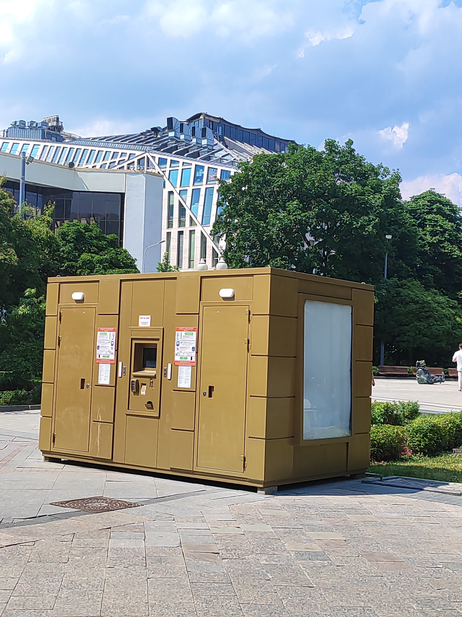 An exterior shot captures a gold-colored, modular kiosk with multiple doors, a central payment or information window, and signage. The kiosk sits on a paved plaza. In the background, a modern building with a distinctive glass roof and facade is visible, along with mature trees. The sky is partly cloudy. A man in a white shirt and light-colored pants walks away from the kiosk towards the right of the frame. Other people and benches are visible in the distance.