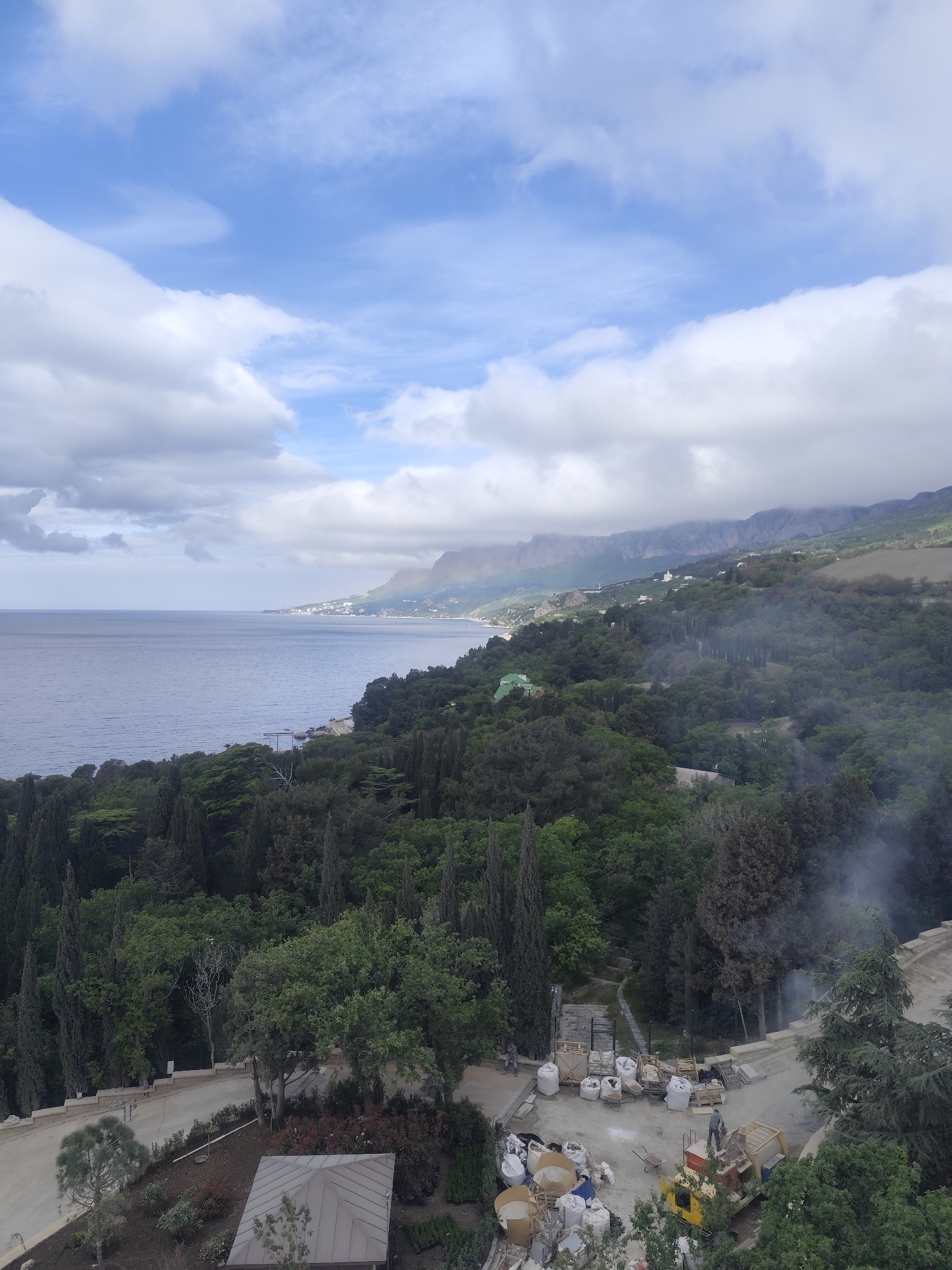 The image shows a coastal scene with mountains in the background under a sky with scattered clouds. The location is Unknown, Unknown. In the foreground, there is lush greenery and trees. A construction or work area is visible, with equipment, materials (such as bags and containers), and a vehicle. There appears to be some smoke or steam rising from the area. The middle ground features more trees leading down to the ocean. The background reveals mountains along the coastline, partially obscured by clouds. The ocean stretches towards the horizon under a partly cloudy sky.