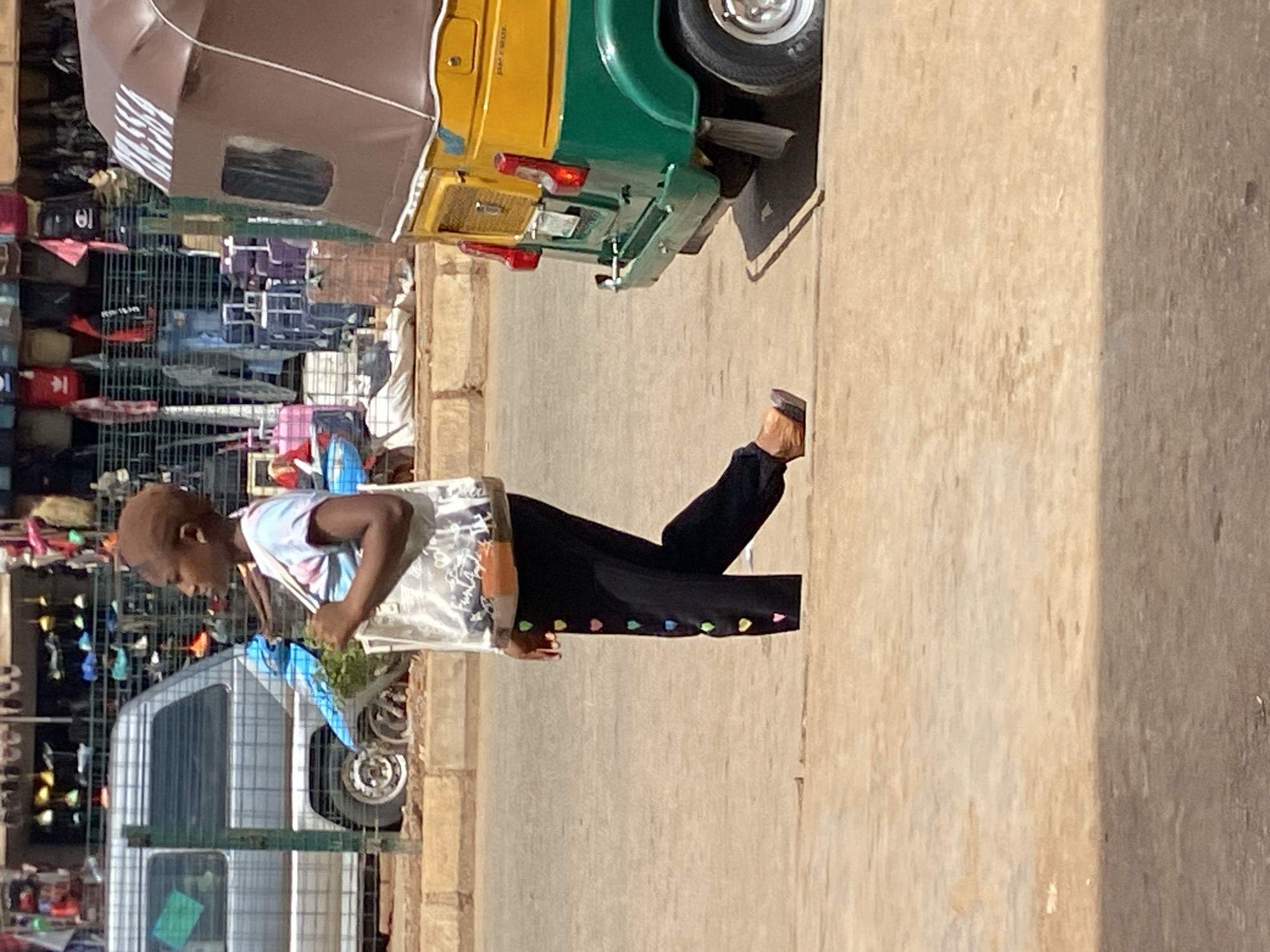 The image, oriented correctly, depicts a bustling street scene in Bukuru, Nigeria, likely during the day.
**Subjects:**
A young individual with short, close-cropped hair is walking on a paved or dirt path on the right side of the frame. They are wearing a light-colored, possibly tie-dye patterned t-shirt and black trousers adorned with a row of small, colorful heart-shaped embellishments near the ankles. They wear dark sandals and carry a translucent or light-colored bag over their right shoulder, which they hold with their right hand. Their gaze is directed downwards or slightly ahead.
**Objects:**
In the background, on the left side of the street, there is a vibrant yellow and green three-wheeled auto-rickshaw (commonly known as a Keke Napep in Nigeria) with a brown canvas canopy, parked or moving slowly. Part of its rear wheel and exhaust pipe are visible. Behind the auto-rickshaw and extending further left are numerous market stalls or racks, enclosed by metal fencing. These stalls display a variety of goods, including shoes, bags, and other items, some hanging and others stacked, indicating a commercial area. Further down the street, partially obscured by the fencing, a white van or minibus can be seen.
**Activity or Event:**
The main activity shown is the individual walking along the street, seemingly navigating through or near a market area. The scene captures a moment of daily life in an outdoor urban or semi-urban environment.
**Notable Details:**
The bright lighting suggests it is daytime, with clear weather. The ground appears to be a mix of dirt and perhaps some paved sections. The type of vehicle and the market setup provide strong contextual cues for a West African street scene. No clearly legible text is visible on any objects or signs. The overall atmosphere appears active and ordinary.