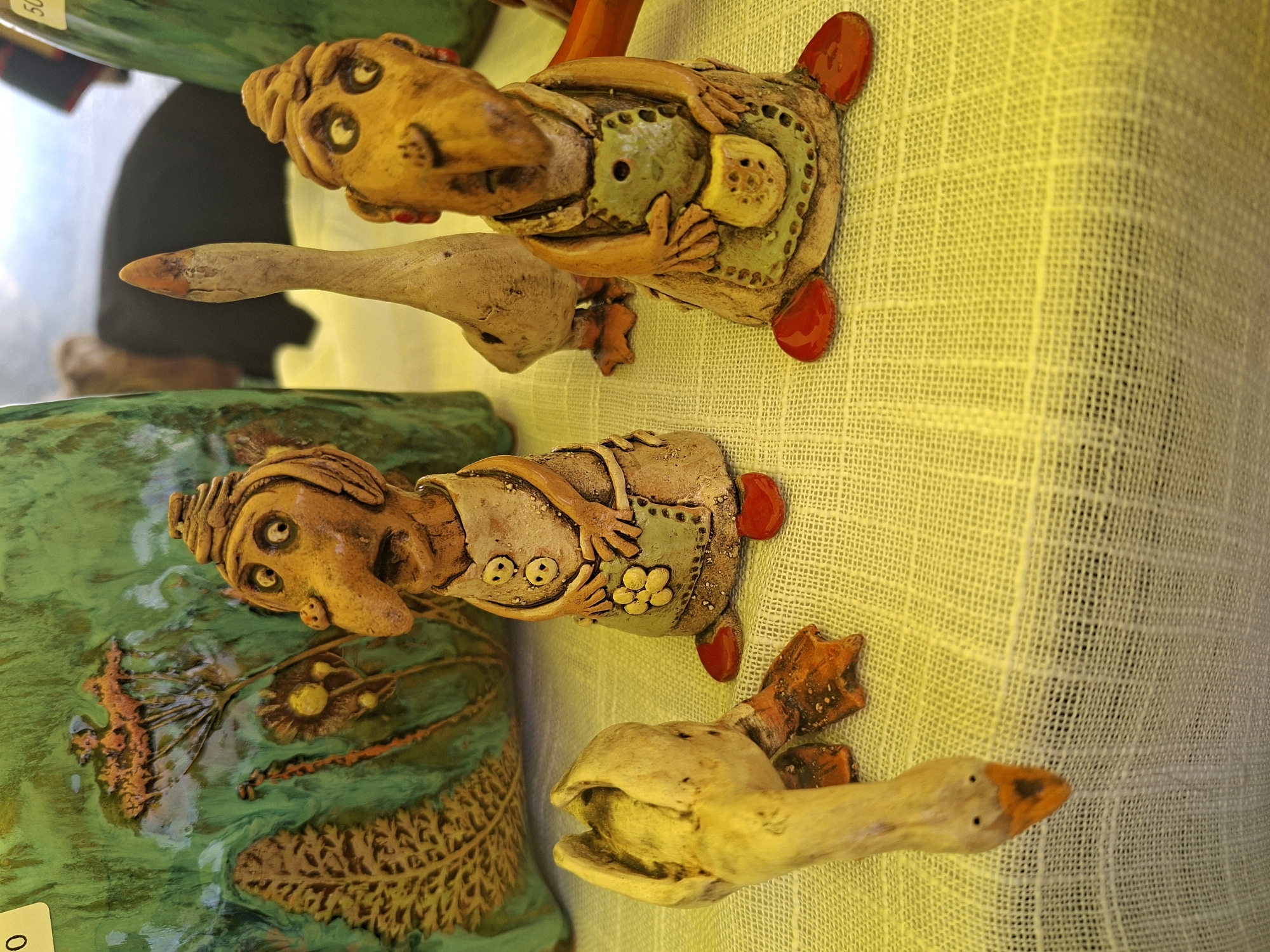 This media file displays a close-up, high-angle shot of three whimsical ceramic figures, possibly decorative geese or ducks with human-like features, arranged on a textured yellow surface, likely a tablecloth or fabric display. The figures are crafted in a folk art style, with expressive, cartoonish faces and simplified body forms.
The top figure is a goose with a long, light-colored neck and head, its beak pointed to the left. The body is yellow, adorned with green and yellow details suggesting clothing or embellishments, and red feet. Its eyes are large and round, giving it a surprised or alert expression.
Below this, a second ceramic figure, also goose-like, rests on a green, textured surface that appears to be part of a larger ceramic piece, possibly a decorative bowl or platter. This figure has a yellow head with prominent, expressive eyes and a beak. Its body is also yellow and features similar decorative elements to the top figure, including what looks like a yellow floral pattern on its chest and red feet.
The third figure, lying in the foreground and partially obscured, is another yellow goose with its beak open, as if honking or calling out. Its wings and feet are visible, also in orange-brown tones.
The overall impression is of charming, handmade crafts, likely presented for sale or display. The lighting suggests it could be daytime, possibly indoors or in a shaded outdoor market. The background is blurred, but hints at other items or displays, common in a retail or craft fair setting. There is a partial view of a green object in the upper left corner with some visible text on a white sticker. A small white tag with the number "0" is visible on the lower left. The scene evokes a sense of rustic charm and playful artistry.