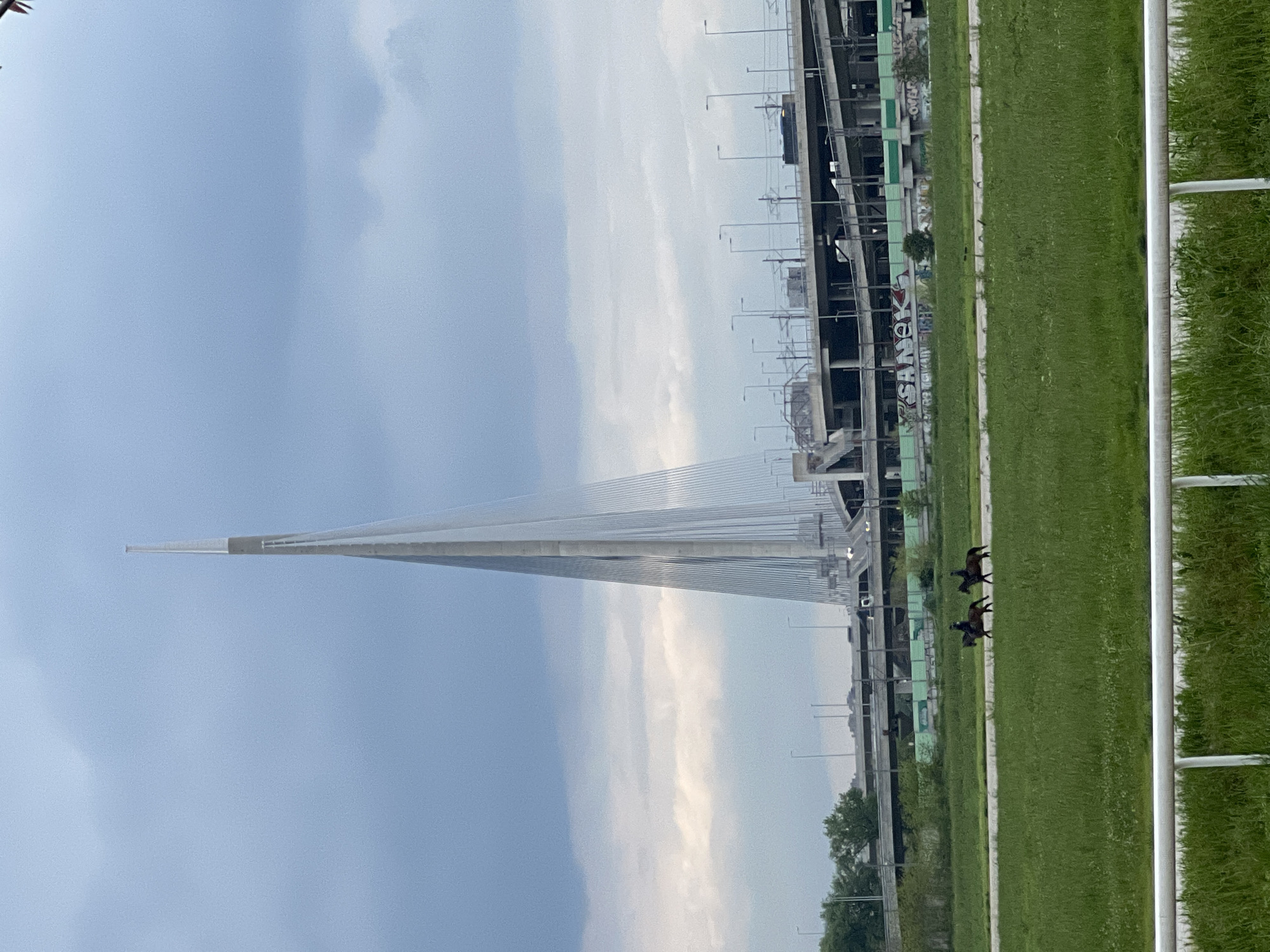 This landscape image, initially rotated 90 degrees clockwise, depicts a modern urban scene in Cukarica Urban Municipality, Serbia, likely near the Ada Bridge and Hippodrome Belgrade.
The central and most prominent feature is a large, white, single-pylon cable-stayed bridge, characteristic of the Ada Bridge, spanning across the frame. Numerous white cables fan out from its tall, slender pylon, supporting the roadway.
In the foreground, a wide, lush green grassy area, possibly a racecourse or training track, stretches horizontally. Two dark horses, each with a jockey in racing silks, are visible on this track, moving from right to left across the frame. A white metal railing separates the viewer's perspective from the grassy field on the far right.
The background reveals a complex urban infrastructure, including elevated roadways, concrete support structures, and what appear to be buildings, some possibly under construction. Streetlights line the bridge and adjacent pathways. On a concrete wall or structure in the background, graffiti is visible, including the words "SANEK" and "OVER" or "OVO".
The sky is overcast with a mix of light blue and soft grey clouds, suggesting diffused daylight, possibly early morning or late afternoon. The overall impression is one of modern infrastructure coexisting with green spaces for equestrian activity.