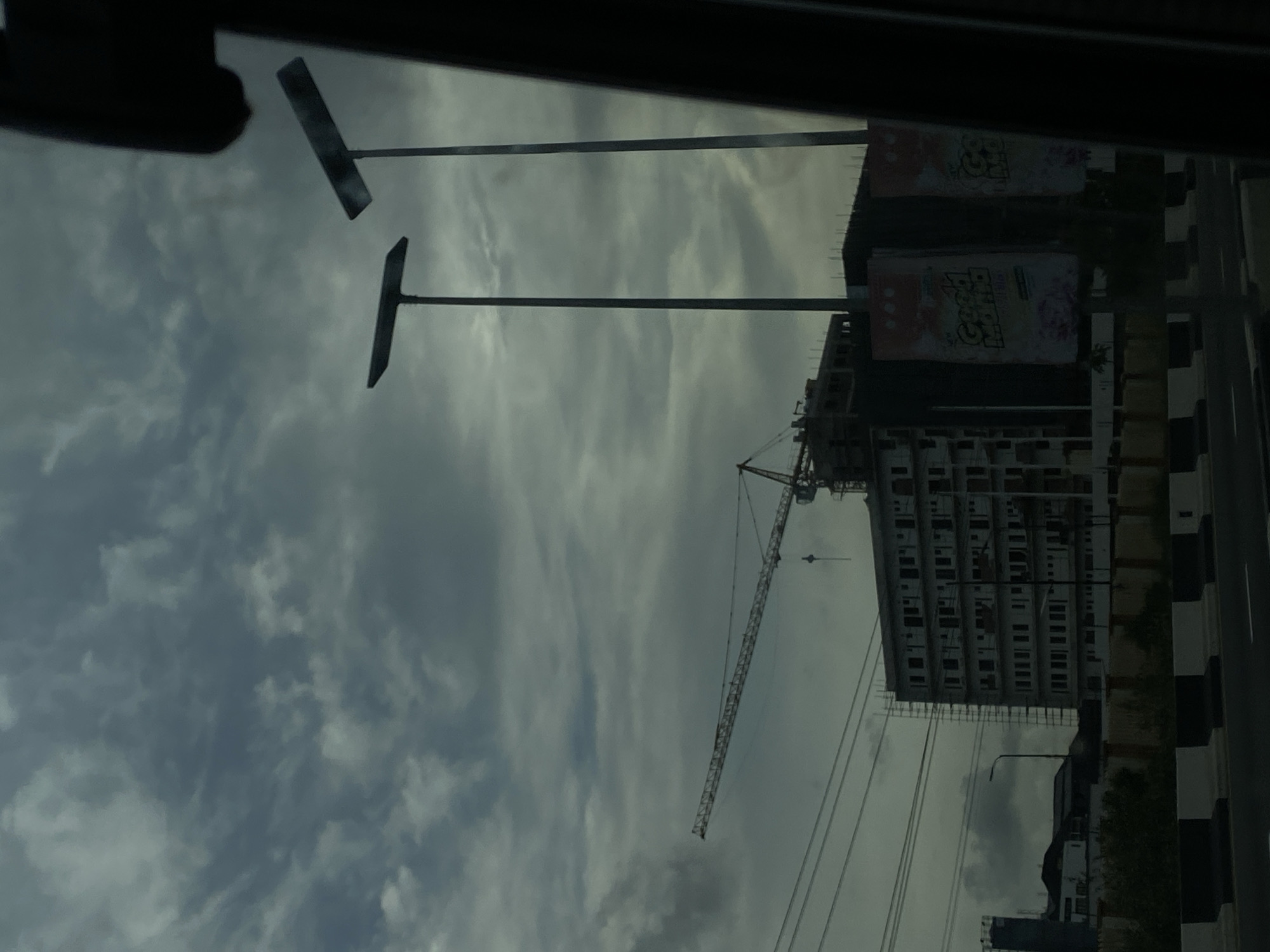 The image, taken from within a vehicle, presents an urban construction scene under an overcast sky. Two dark, T-shaped structures, possibly streetlights or signs, are prominently silhouetted against the dense, grey clouds in the upper left.
On the right side of the frame, a multi-story building is visibly under construction, featuring exposed concrete floors and an unfinished facade. A large tower crane extends from behind and above this building, with its long jib spanning across the mid-ground. Attached to the side of the unfinished building are large banners; one clearly displays the text "Grand Opening," while other promotional text is partially visible.
Along the far right, a distinct black and white striped curb or road barrier marks the edge of a street. Utility lines are also visible, stretching horizontally across the scene. The overall lighting is diffused due to the heavy cloud cover, indicating a cloudy day rather than a specific time like dawn or dusk. The presence of significant construction activity suggests ongoing urban development, consistent with a dynamic city like Lagos, Nigeria.