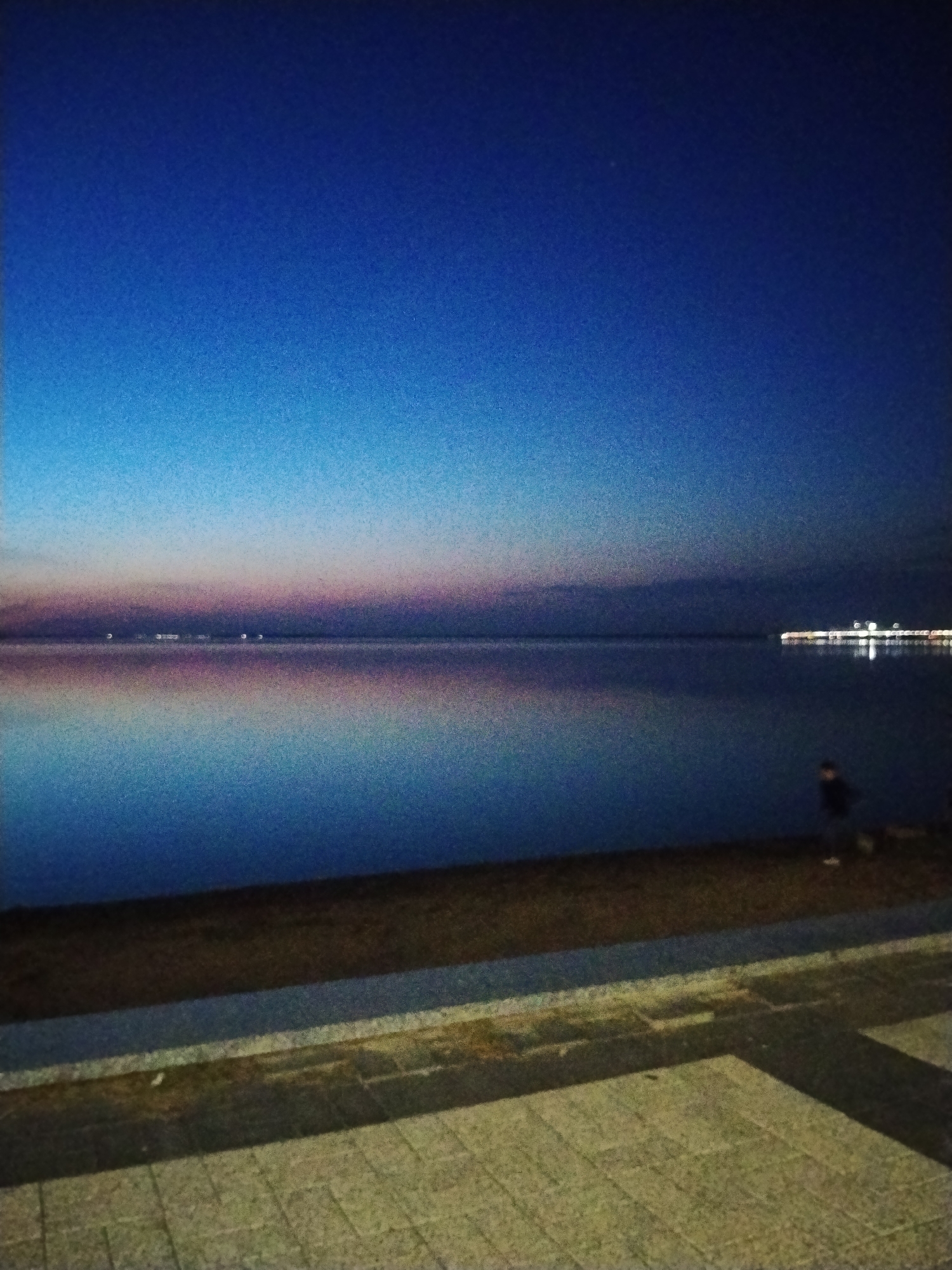 The image captures a twilight scene over a body of water, likely Lake Burabay in Kokshetau, Kazakhstan, given the context of the prompt. The sky transitions from a deep blue at the top to lighter hues of blue and pink near the horizon. The water is calm, reflecting the colors of the sky and distant lights. On the right side of the image, a pier with several lights extends into the water, creating reflections on its surface. In the foreground, a person, appearing to be a child due to their small stature, is visible on the shore, silhouetted against the water. The shore is a strip of dark earth, transitioning into a paved walkway made of grey and beige rectangular stones. The overall atmosphere is serene and peaceful.