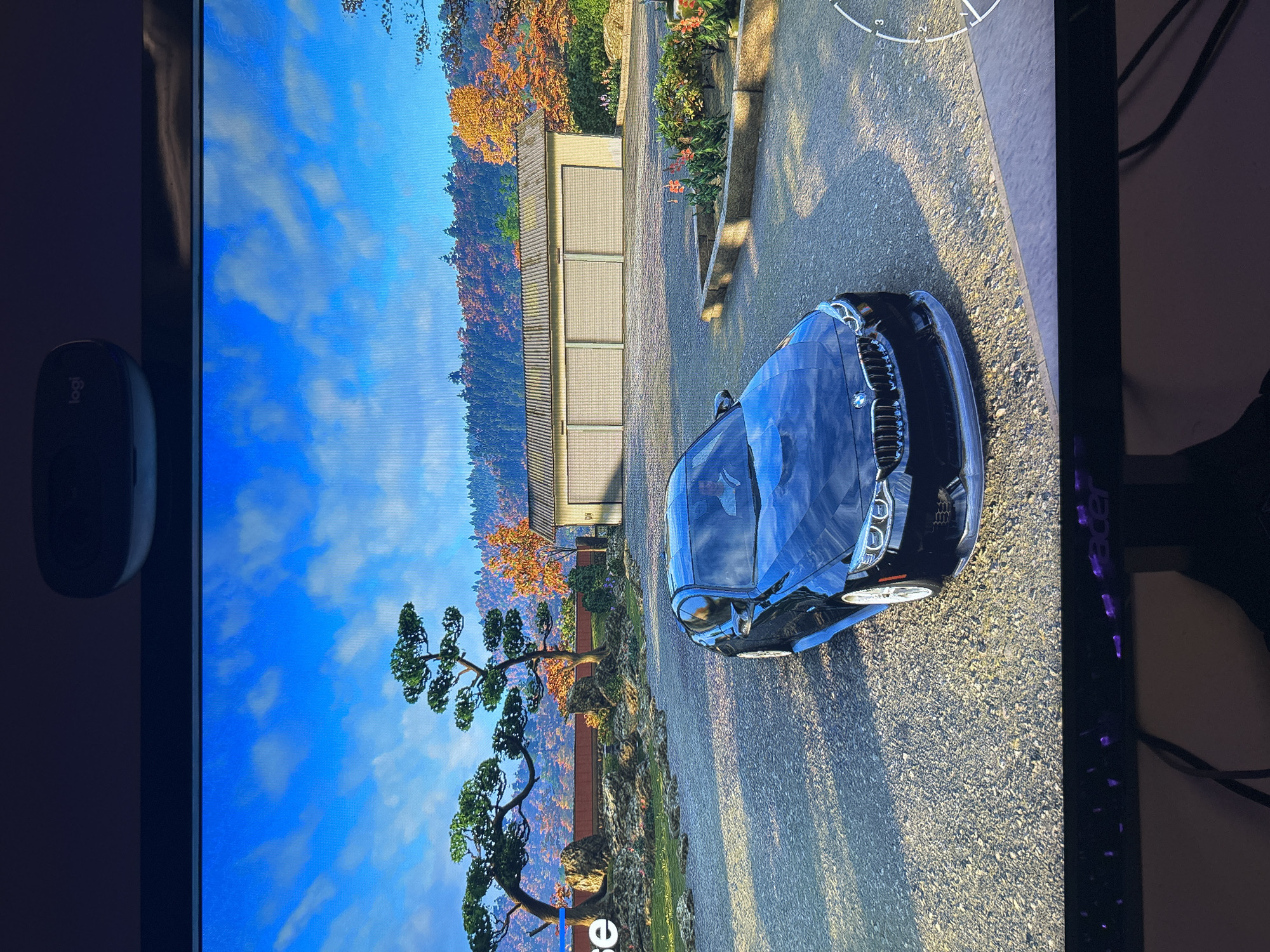The image shows a screen displaying a video game, possibly Forza Horizon 5, set in a visually detailed environment. A dark blue BMW car is parked on a cobblestone or asphalt road, casting a long shadow. The environment suggests a residential area or a scenic drive in Japan, featuring traditional Japanese-style buildings with multiple garage doors, meticulously landscaped trees (bonsai and maples), and a backdrop of forested hills transitioning to autumn foliage. The sky is blue with scattered clouds.
The presence of a Logitech mouse to the left of the screen and a partially visible monitor (Acer) with illuminated purple lights on the right indicates the scene is captured from a personal computer setup.