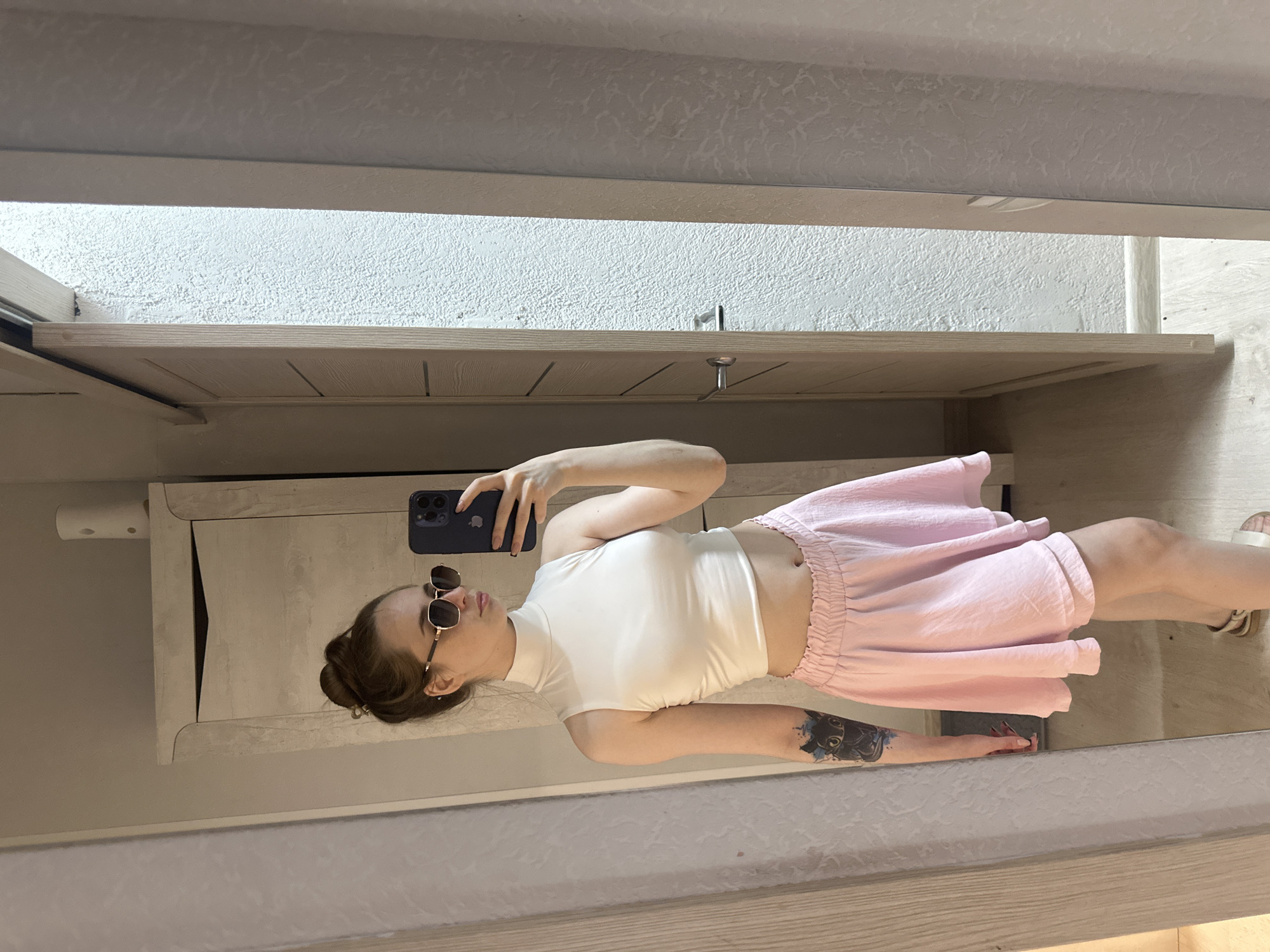 A young woman is taking a mirror selfie in a room. She is wearing a white sleeveless turtleneck crop top and a light pink ruffled skirt. She has her hair pulled up in a bun and is wearing sunglasses. She has a tattoo of a black cat on her left arm. The room appears to be a bedroom or dressing area, with a large mirrored closet and a closed door visible in the background. The flooring is light-colored wood.
