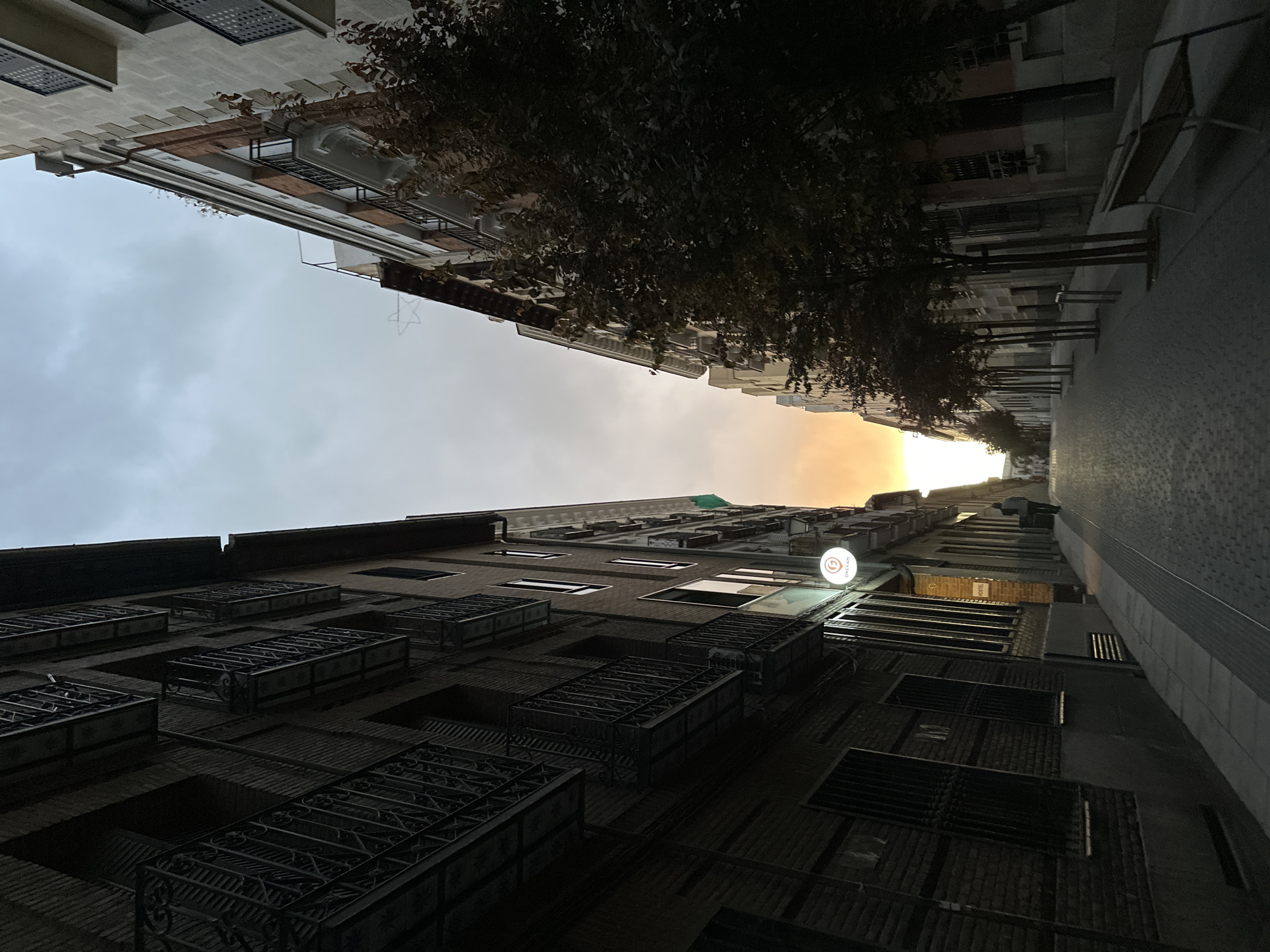 The image depicts an upward view of a street in Madrid, Spain. The perspective emphasizes the buildings lining the narrow street, with the sky visible between them. The architectural style of the buildings is consistent with that found in Madrid.
Details:
* **Buildings:** The buildings are multi-story structures with balconies and windows.
* **Sky:** The sky is visible between the buildings, with the sun appearing to create a bright illumination.
* **Street:** The street appears to be paved.
* **Trees:** There are some trees along the street.
* **Lighting:** The natural lighting suggests either a sunset or sunrise.
* **Sidewalk**: A sidewalk is seen on the right.
* **Bench**: A bench is seen on the right.
* **Sign**: There is a white sign with a logo on one of the buildings.