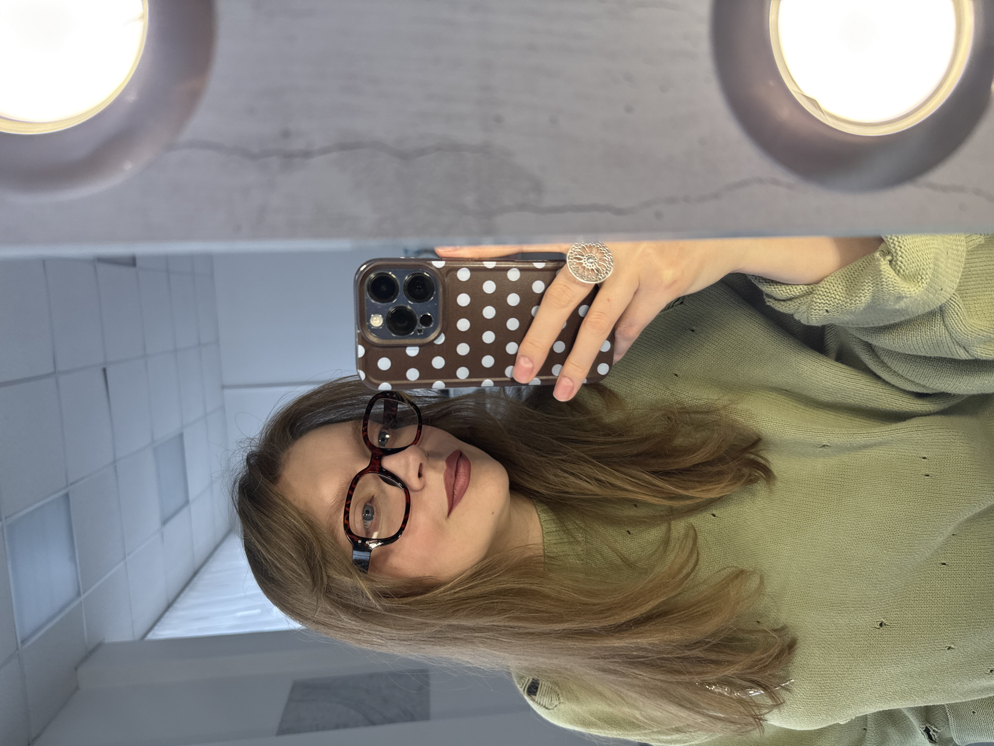 A woman takes a selfie in a bathroom. She wears glasses, a green sweater, and a large ring. She is holding a brown phone with white polka dots. The bathroom has white tiles and circular lights on the mirror. There is no evidence to suggest that this scene is located in Izhevsk, Russia.
