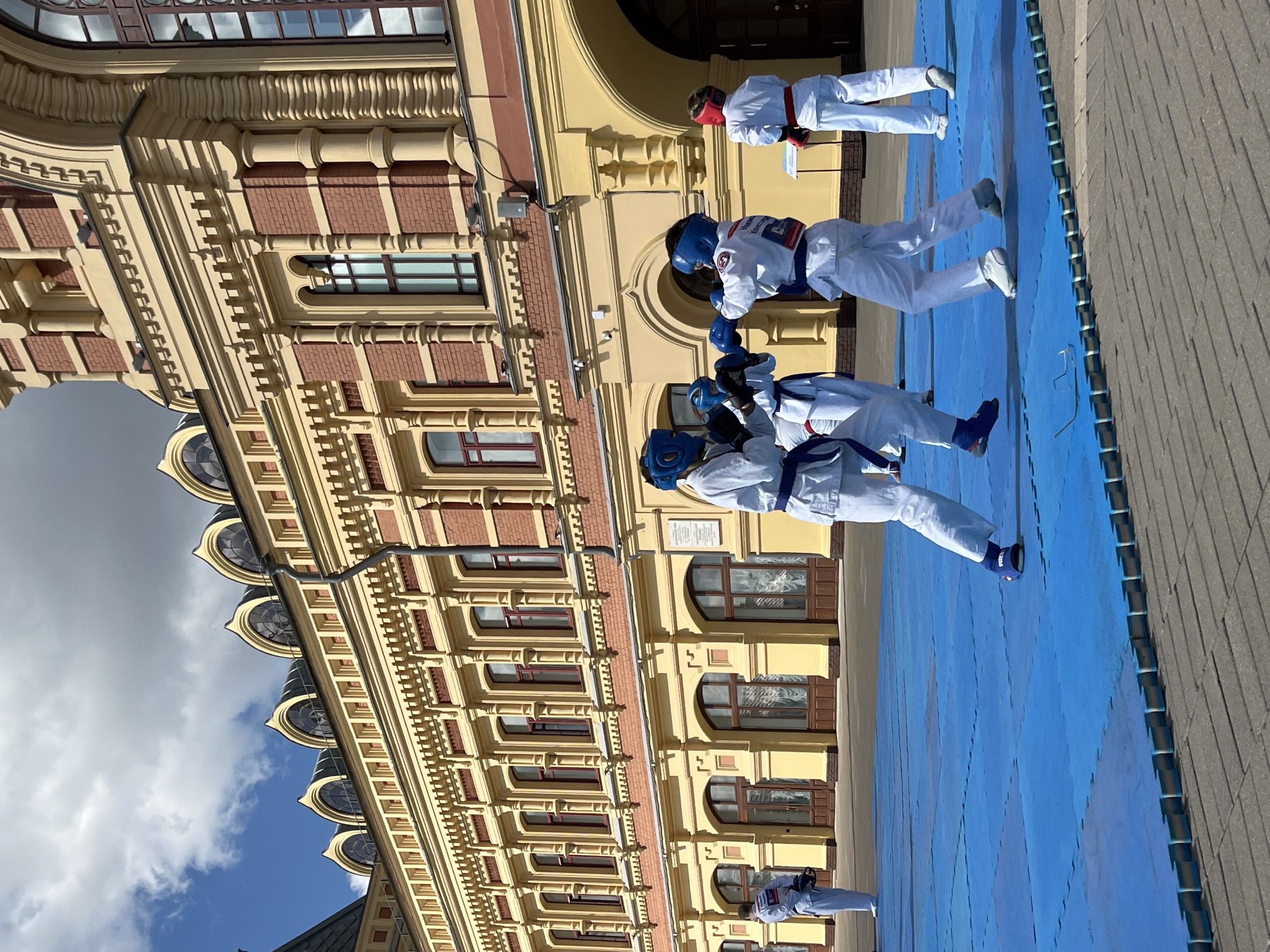 The image captures a training session for young martial artists, likely a form of karate or taekwondo, outdoors in front of a grand, yellow-and-red brick building with ornate architectural details, including arched windows and decorative cornices. The training takes place on a blue matting surface.
In the foreground, two individuals in white gis and blue headgear are engaged in sparring, with one performing a punch towards the other. Both wear blue belts. To the left, another individual in a white gi and blue belt stands facing away from the camera, holding protective headgear. To the right, a fourth individual in a white gi and red belt faces the sparring pair, their back to the camera.
The building appears to be a significant historical or institutional structure, possibly an educational institution or a museum, given its elaborate facade. The sky is partly cloudy, suggesting daytime. The overall setting conveys a sense of organized activity in a formal, urban environment. The building's architectural style is indicative of 19th-century Russian Revival or Eclectic architecture.
This building is identified as the Nizhny Novgorod State Academic Drama Theater named after Maxim Gorky, located in Nizhny Novgorod, Russia. The theater is a prominent landmark in the city's historical center.
