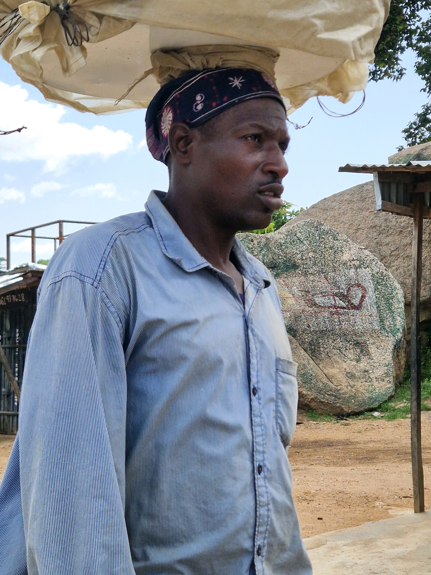 A man is shown from the chest up, looking to his right. He has a dark complexion and is wearing a blue striped collared shirt with buttons. On his head, he carries a large, light-colored fabric bundle. He is wearing a dark bandana with a patterned design. Behind him, a large, textured rock is visible, with what appears to be a red drawing on its surface. To the right of the rock, a portion of a simple wooden structure with a corrugated metal roof is present. The ground is sandy and there are some green plants. The sky is visible above with some clouds. A metal fence structure is partially visible on the left side of the image.
