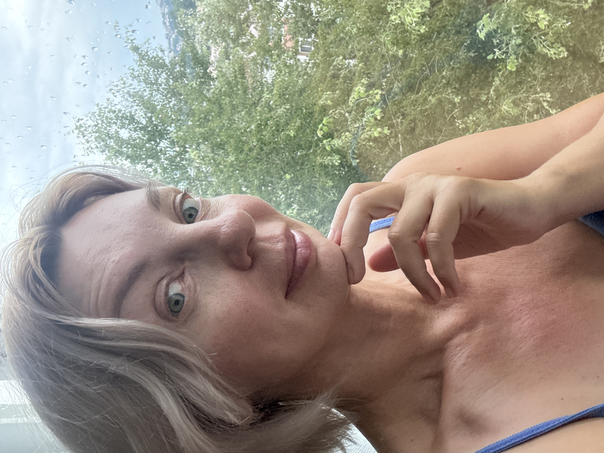 A middle-aged Caucasian woman with short, blonde hair is positioned in the foreground, facing slightly to the right with her chin resting on her hand. She is wearing a blue top. Behind her, a window displays raindrops on the glass, with lush green trees and a few distant buildings visible through it. The overall setting appears to be an urban environment, possibly Sochi, Russia, indicated by the presence of trees and buildings in the background. The lighting suggests an overcast or rainy day.