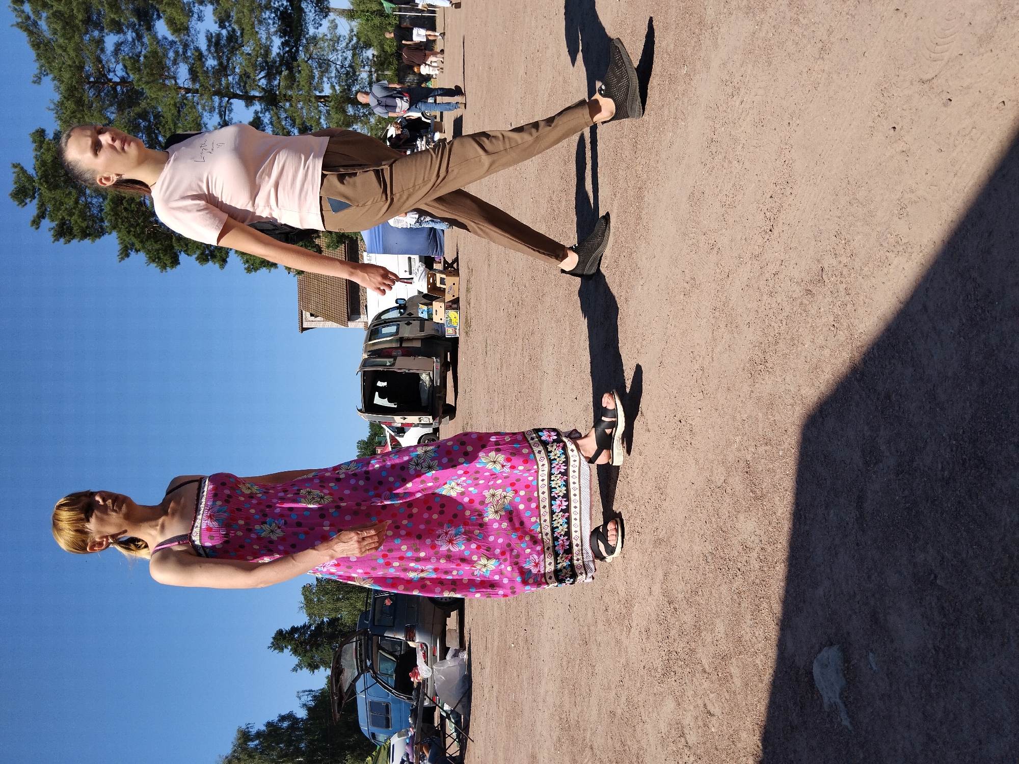 The image depicts an outdoor scene, possibly a market or fairground, with a sandy ground surface. Two women are the primary subjects. The woman on the left wears a long, bright pink dress with a floral pattern of blue, yellow, and pink circles and flowers. She has blonde hair pulled back in a ponytail and wears dark sandals. The woman on the right is dressed in a pale pink t-shirt and brown trousers. She has dark hair pulled back in a ponytail and wears black sneakers. Her gaze is directed forward, while the other woman looks slightly to her left. In the background, several vehicles, including vans, are parked. Some people, appearing to be vendors or shoppers, are visible near the vehicles and a blue tent. The overall environment suggests a sunny day, with trees and a building visible in the distance. The presence of parked vehicles and tents hints at a temporary event setup.