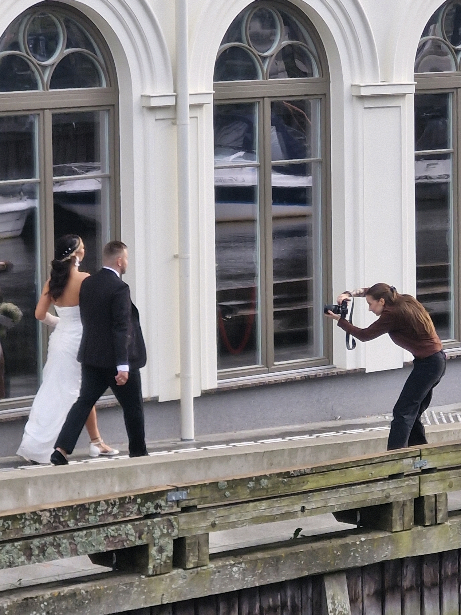 A medium shot captures a wedding couple walking from left to right, with a photographer capturing their moment. The bride, on the left, wears a white strapless wedding gown and her dark hair is styled with white flowers. The groom, to her right, is in a black suit with a white shirt and tie. A woman, the photographer, stands to the right of the couple, facing them and holding a black camera with a strap around her neck. She is dressed in a brown long-sleeved shirt and black pants, crouching slightly as she takes a photo. The backdrop features a white building with arched windows. The scene is set outdoors, with a wooden structure in the foreground.