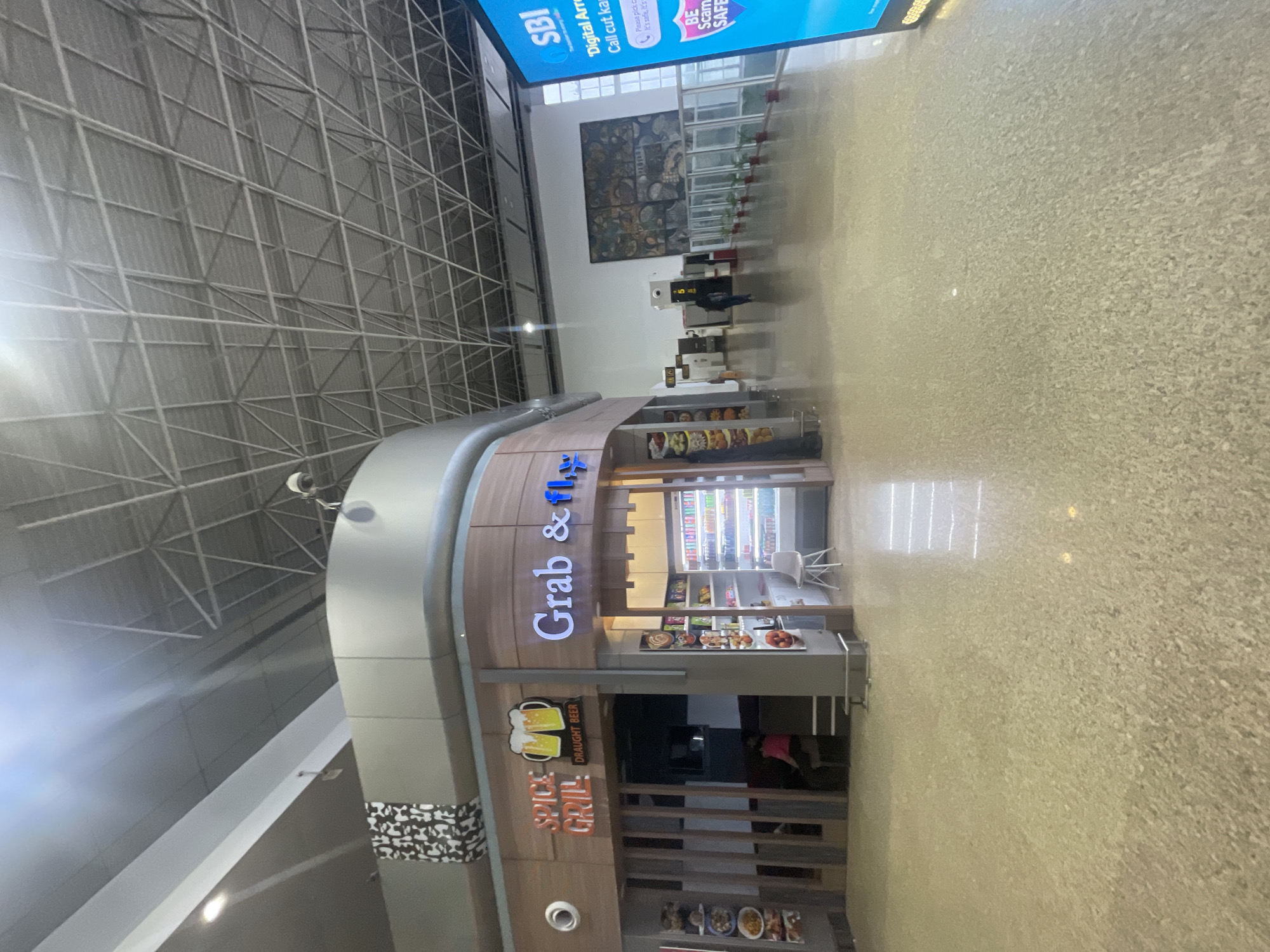 This image shows the interior of an airport terminal in Raipur Khurd, India. In the foreground, a polished floor reflects the overhead lighting. To the left is a commercial establishment named "Spice Grill" which appears to be a restaurant or bar, with signage featuring images of beer mugs and the text "DRAUGHT BEER". Adjacent to it, to the right, is a "Grab & Fly" convenience store with a brightly lit display of drinks and snacks. In the midground, there is a wide walkway with several potted plants lining the right side. A large mural or artwork adorns the wall in the distance. Further back, there are check-in counters and airport signage, including a large digital advertisement for SBI. A few individuals are visible in the background, one walking down the concourse and another sitting inside the Spice Grill. The ceiling is a complex structure of metal beams and panels, typical of a modern airport.