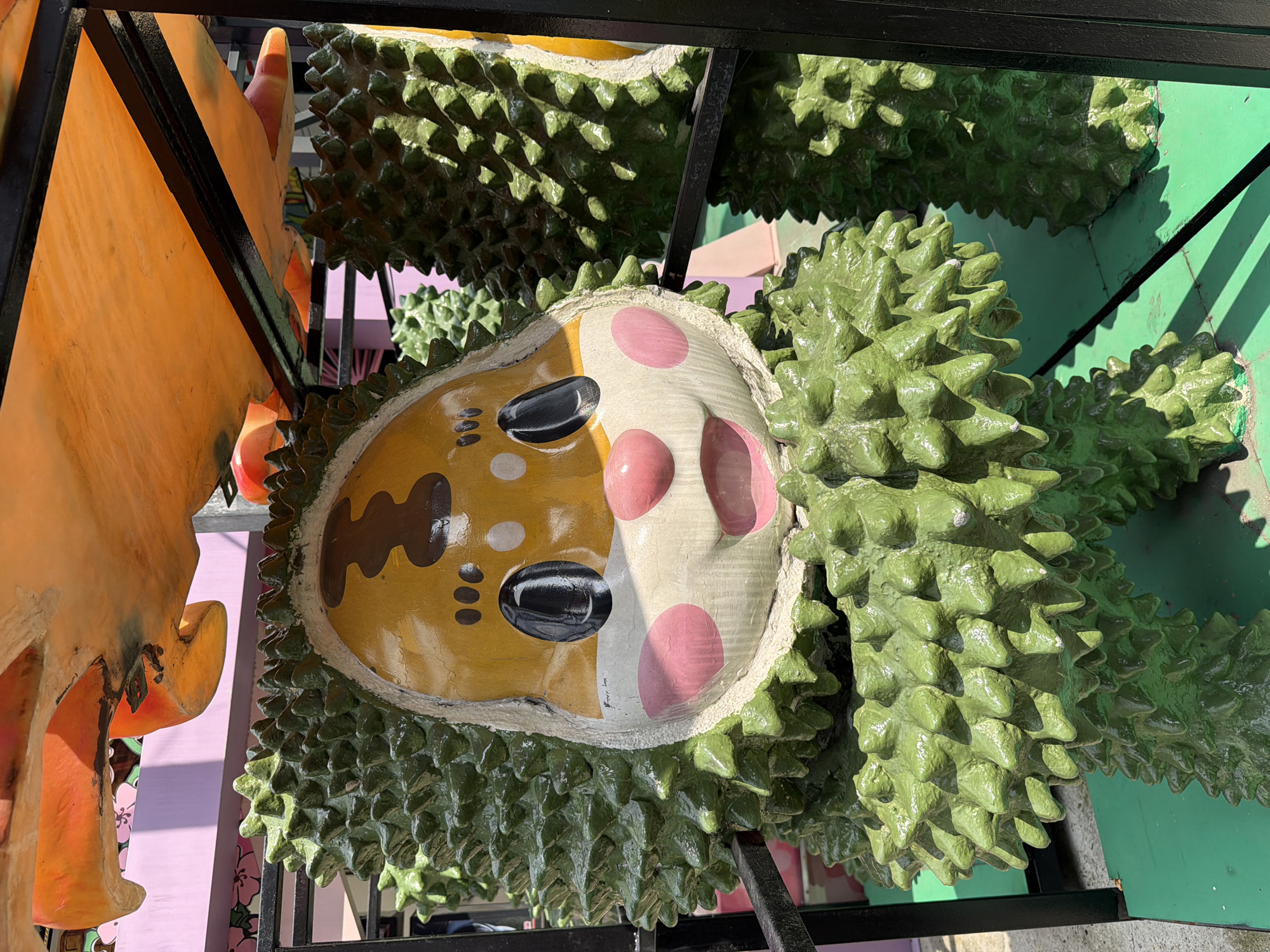 This media file features a close-up, angled view of a decorative sculpture of a durian fruit with a human-like face. The sculpture is predominantly green with a textured, spiky exterior, characteristic of a durian. The face is painted with a yellow base, featuring black eyes, brown eyebrows, and pink circles on the cheeks. There are also three small black dots below the eyes, possibly representing freckles or a smile.
The sculpture appears to be part of an outdoor display, possibly in Kuala Lumpur, Malaysia, given the location context. The lighting suggests it is daytime, with harsh shadows indicating bright sunlight. The background is blurred but shows hints of a green wall and a purple surface, along with structural elements that suggest a stand or frame holding the sculpture. An orange, abstract shape is visible on the left, possibly another decorative element or part of a larger installation.
The overall impression is of a whimsical and perhaps slightly kitschy display, likely intended for a tourist attraction or a festival celebrating local produce like durian. There is no visible text in the image. The focus is on the detailed rendering of the durian sculpture's texture and its anthropomorphic facial features.