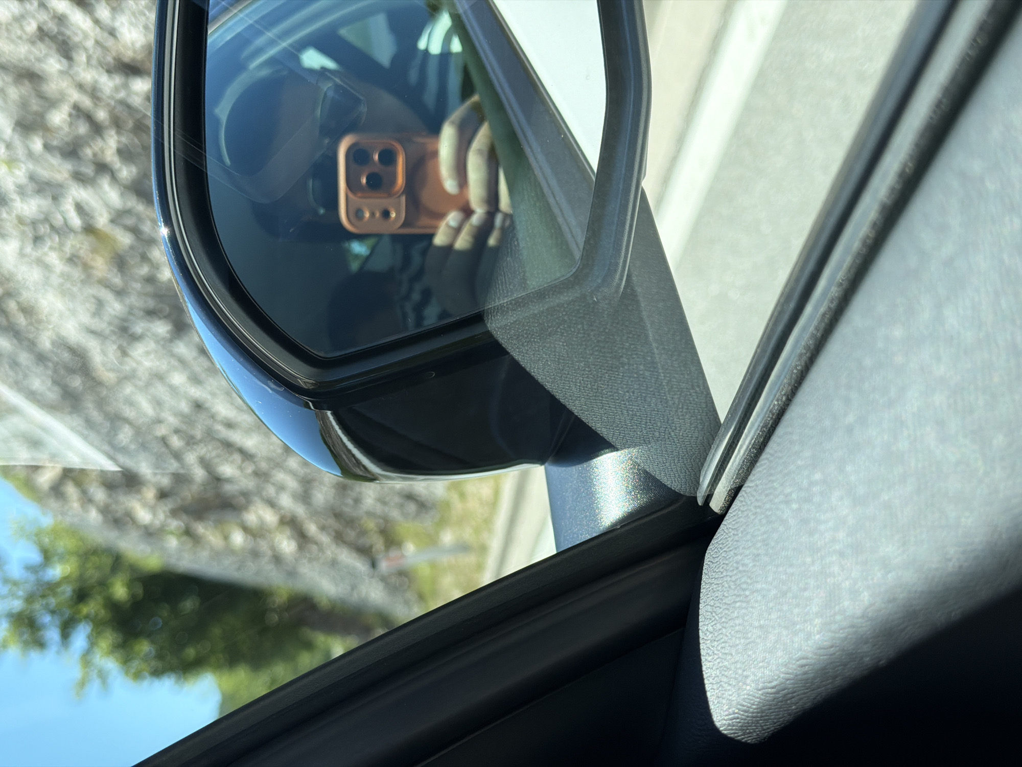 The image is a reflection in a car's side mirror, showing a person holding a smartphone. The smartphone has a prominent orange case with multiple camera lenses. The person is wearing a striped shirt and sunglasses, partially obscured by the reflection. Outside the car, a rocky hillside and green foliage are visible under a bright blue sky. The scene appears to be captured from within a moving vehicle, suggested by the angle and the slight blur of the background. The location is identified as Vrela, Montenegro.
