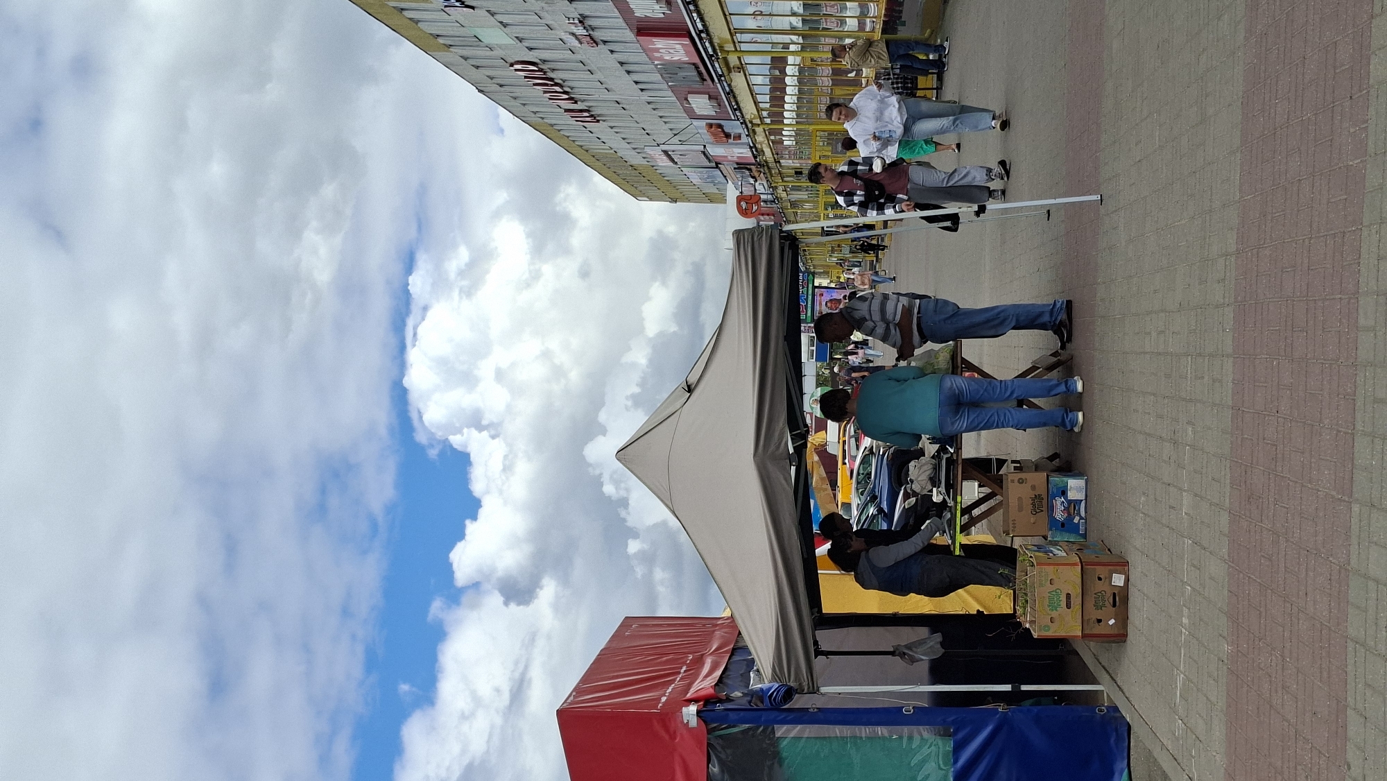 The image depicts an outdoor market scene in Maladzyechna, Belarus. In the foreground, a temporary stall with a beige canopy is set up, displaying various items on a table. Three individuals are gathered around the table, appearing to be interacting with the merchandise. To the left of the stall, a larger, partially visible red and blue tent structure is present. In the background, a long, multi-story building with numerous windows and signage lines the street. Several cars are parked along the side of the building. Other pedestrians are visible walking along the paved pathway in the middle ground and background. The sky is partly cloudy with a mix of blue sky and white clouds. The ground is paved with interlocking bricks in alternating shades of grey and red. The scene suggests a typical market day with vendors and shoppers.