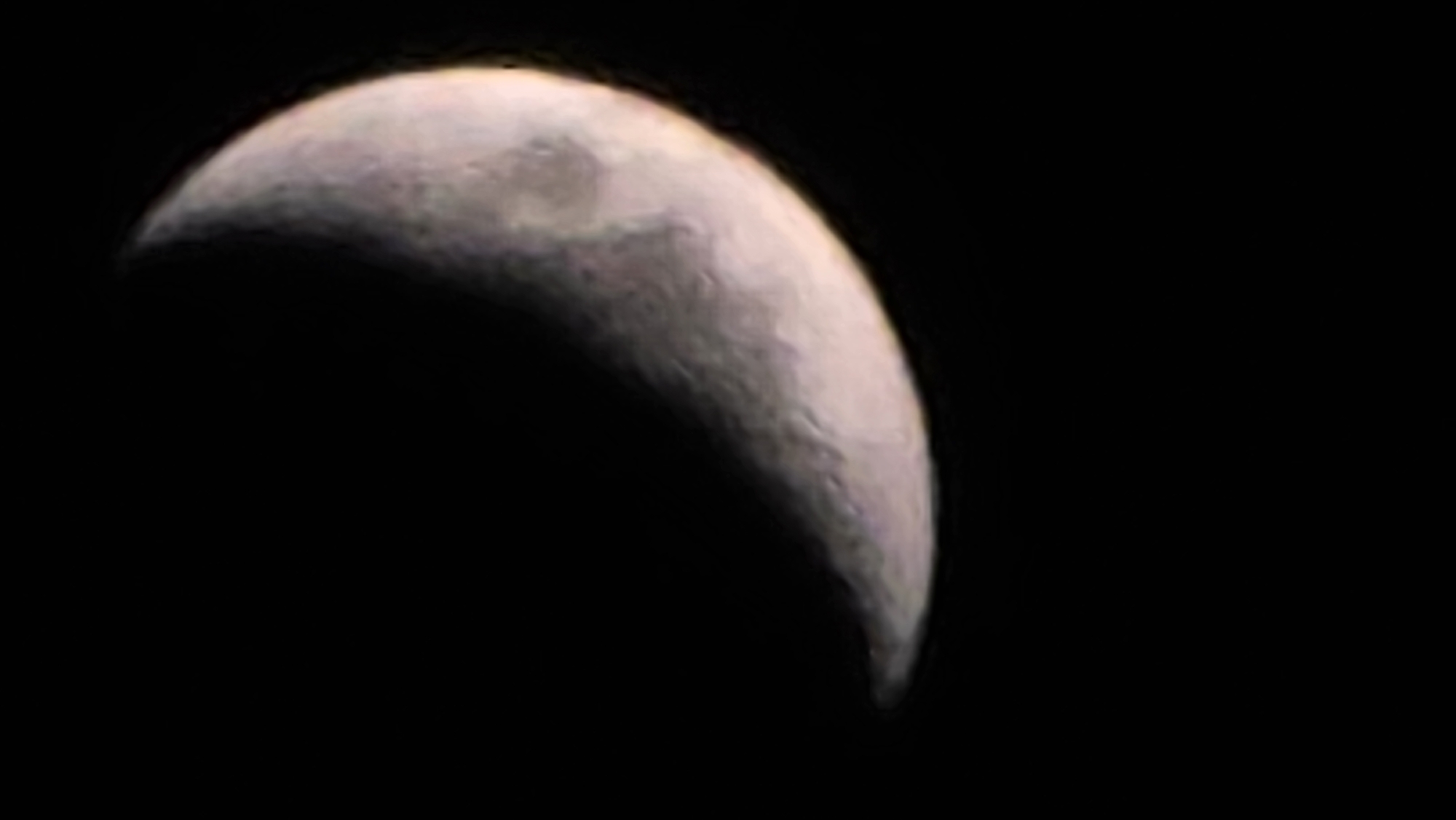 The image is a close-up photograph of a crescent moon against a black background. The moon is partially illuminated, showing its curved edge and some surface details. The colors of the moon appear to be shades of white, gray, and hints of pink and yellow, suggesting atmospheric effects or color processing. There is no indication of the city of Blagoevgrad, Bulgaria, or any other terrestrial environment. The scene is solely focused on the celestial body.
