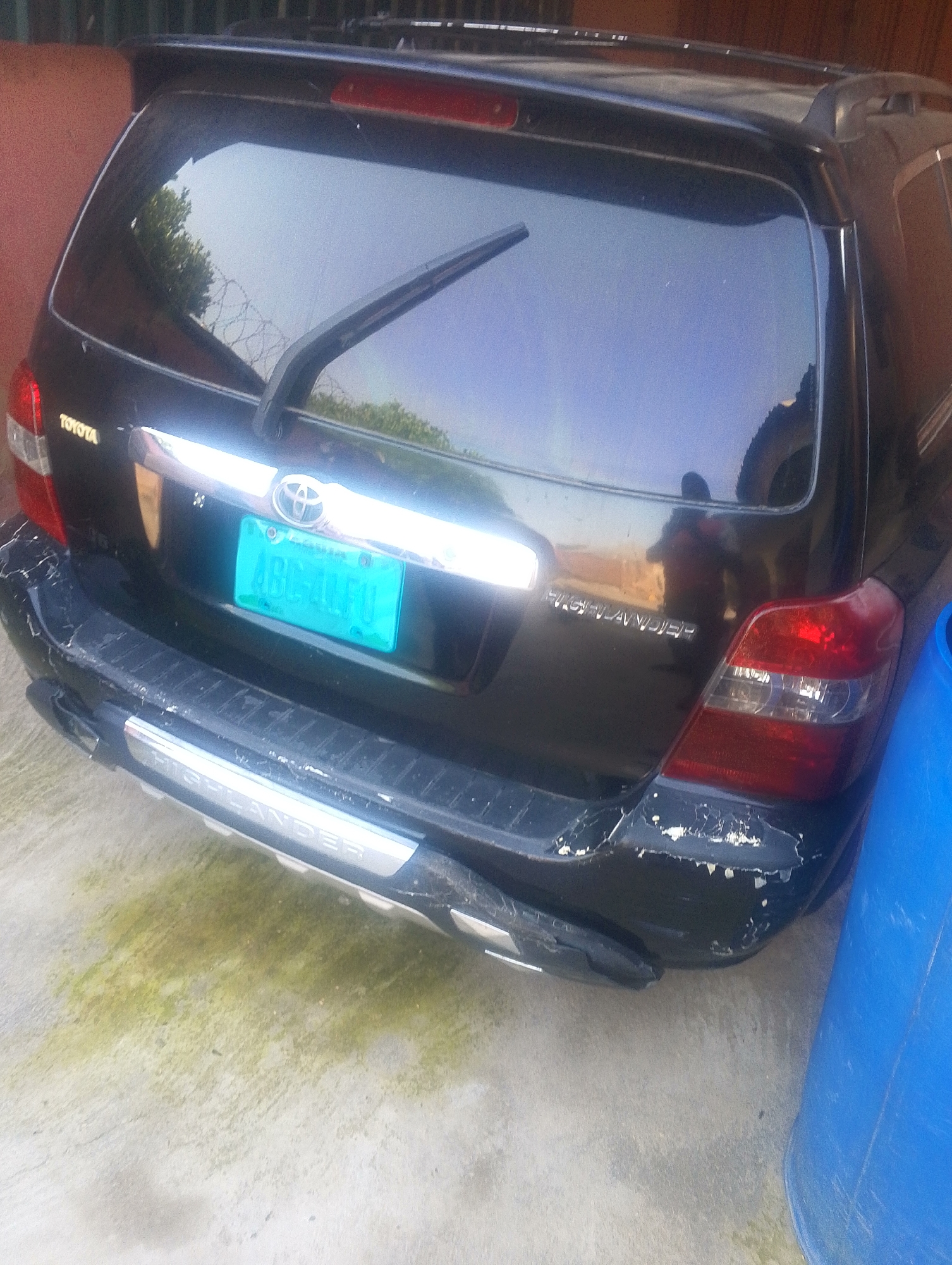 The image shows the rear of a black Toyota Highlander, parked on a concrete surface with patches of moss. The vehicle has a light blue license plate with the characters "ABC-40FU". The rear bumper shows signs of damage, with paint chipped and scraped off in several areas. A large blue plastic container is visible to the right of the vehicle. The scene appears to be outdoors, possibly in a residential or commercial area in Jalingo, Nigeria, given the visual context of the environment. The overall impression is of a functional but aging vehicle in an everyday setting.