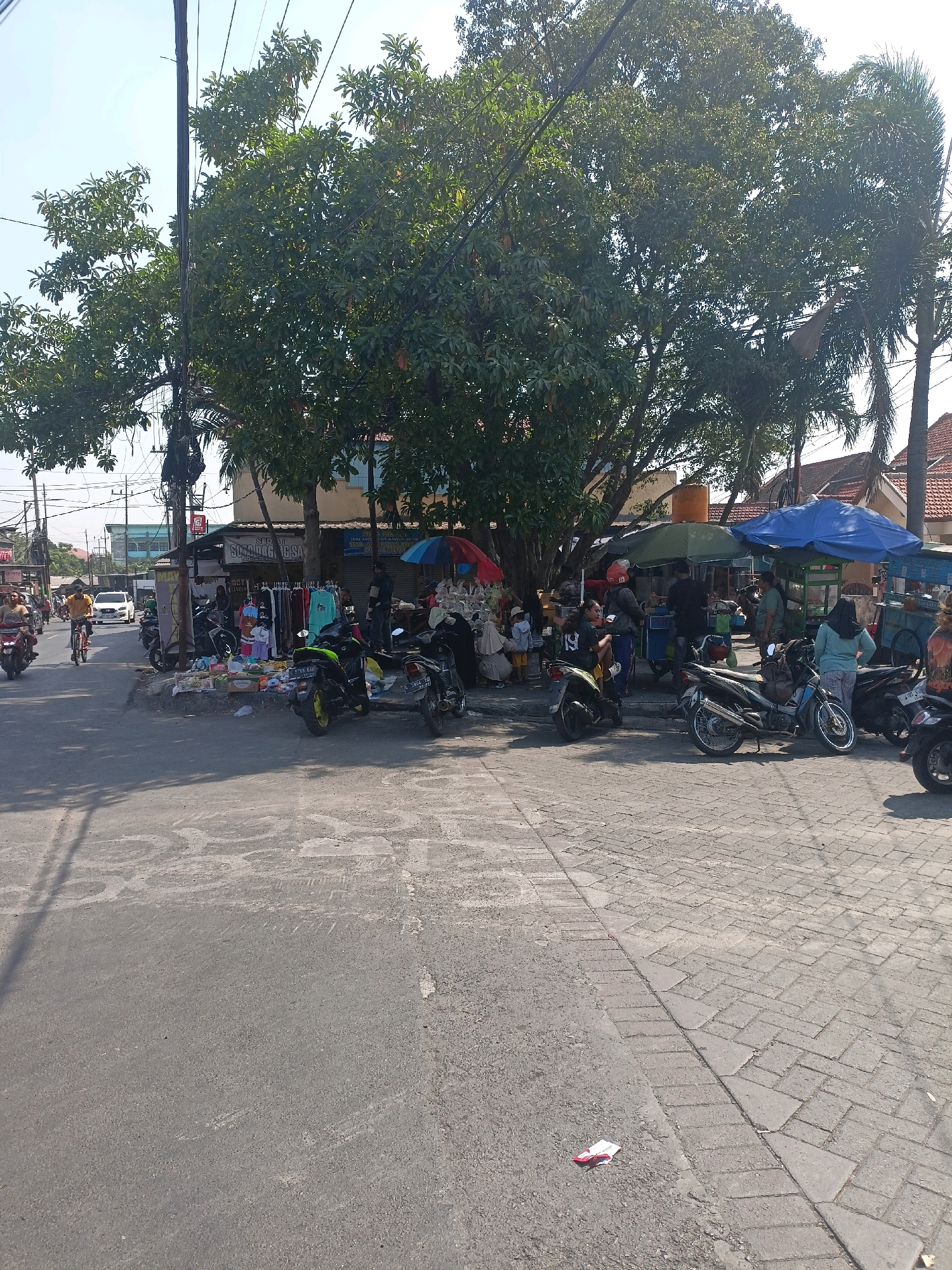 The image depicts a busy street scene in Surabaya, Indonesia, under a clear sky. In the foreground, a paved street transitions to a brick sidewalk on the right. Several motorbikes are parked or in motion on both the street and sidewalk. Along the sidewalk, a marketplace is set up with vendors selling various goods from stalls and carts, shaded by umbrellas and trees. People are browsing the merchandise, interacting with vendors, and moving about. A variety of clothing items are displayed on racks. Buildings with tiled roofs are visible in the background, along with power lines and poles. The atmosphere suggests a typical day in a vibrant urban commercial area.