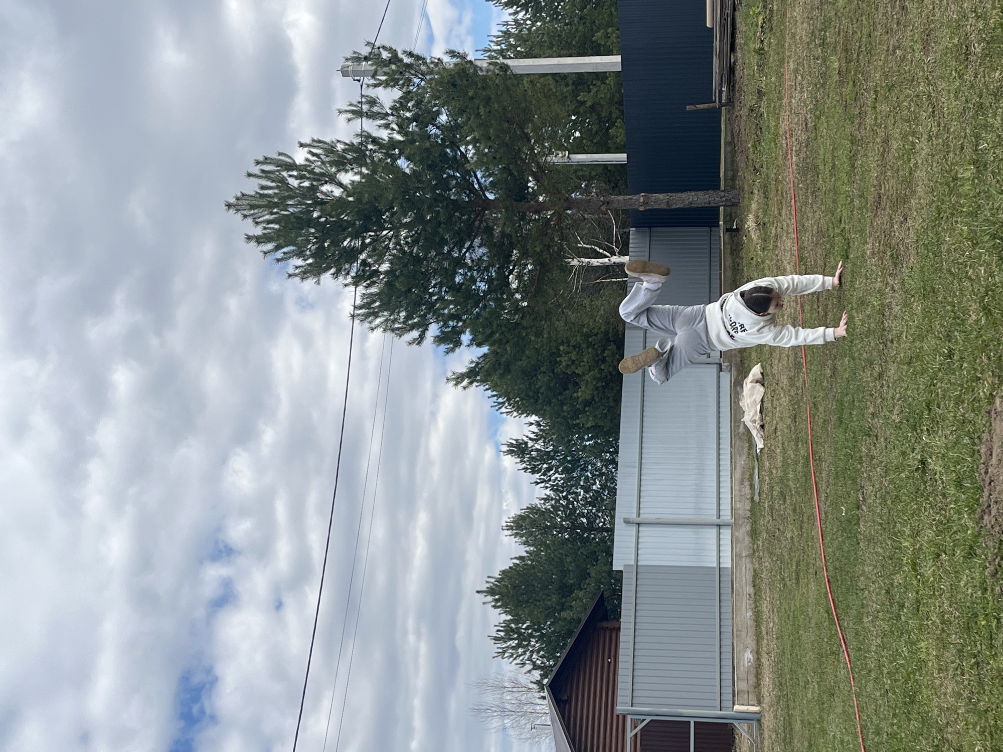 A person wearing a white hooded sweatshirt and gray sweatpants is captured mid-movement, appearing to be in a handstand position, on a grassy lawn in what seems to be Гаревая, Russia. A red hose lies on the grass. Behind the person is a gray metal fence, a pine tree, and parts of a house with a dark brown roof. A section of a dark blue fence is also visible. Overhead, a sky with white and gray clouds is present, alongside power lines.