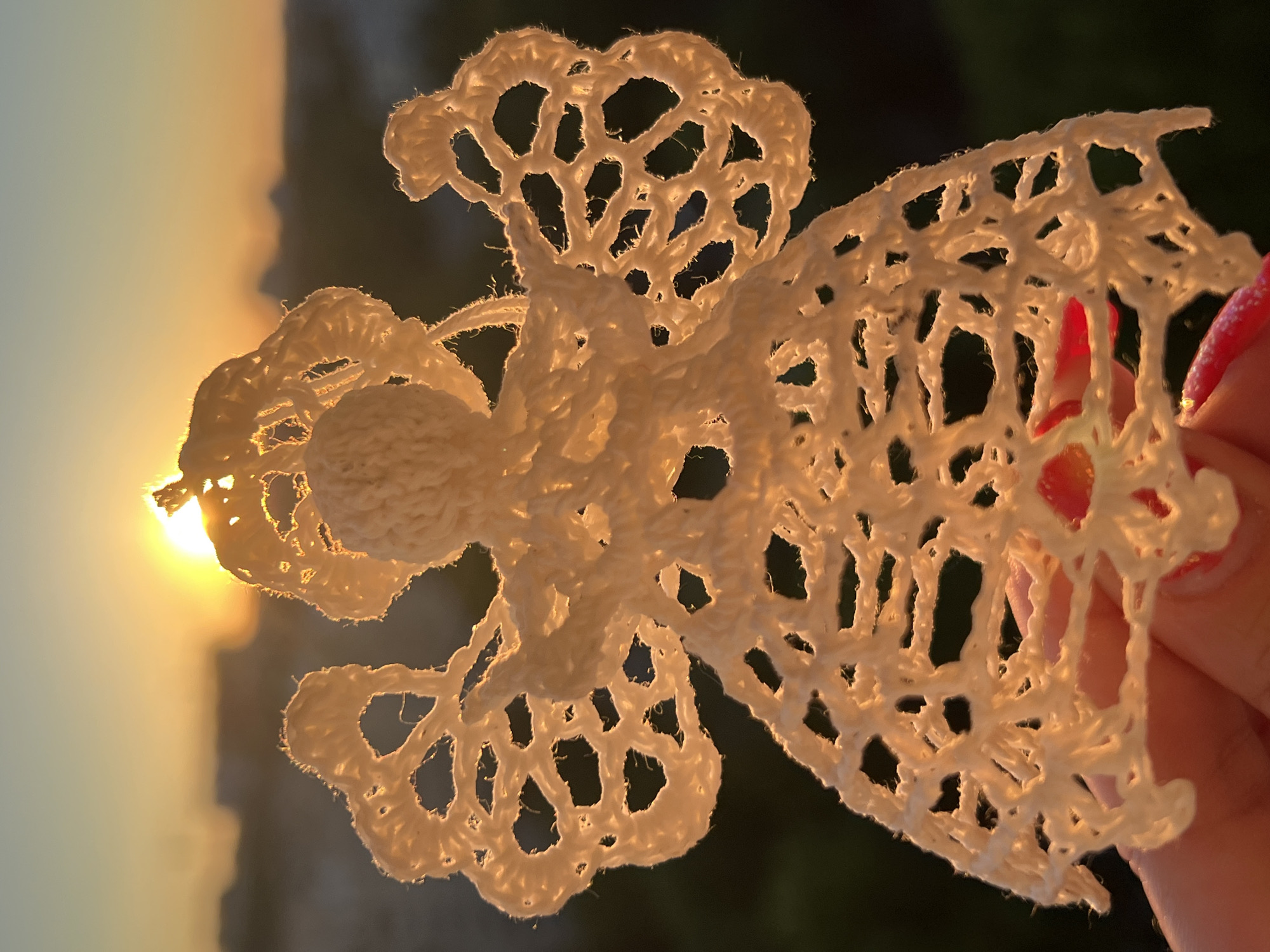 A crocheted white angel ornament is held up against a golden sunset with a hazy cityscape in the background. The sun is partially obscured by the top of the angel's head. The angel's wings and dress are made of open, lacy crochet work, allowing the sunset light to shine through. A human hand with red-painted fingernails is visible at the bottom of the frame, supporting the angel. The background shows a dark, out-of-focus silhouette of buildings, suggesting an urban setting. The overall lighting is warm and golden, characteristic of sunset.