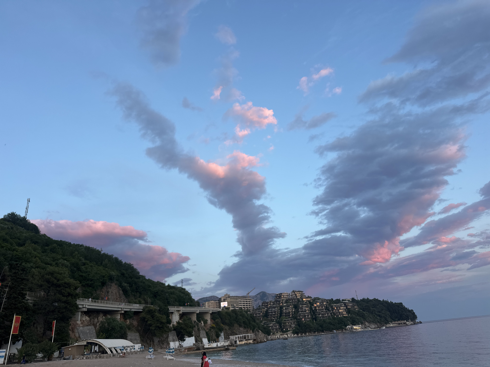 The image shows a beach in Budva, Montenegro, at dusk. The sky is filled with clouds colored pink and gray by the setting sun. A bridge spans the green hillside to the left, while multistory buildings line the coast to the right. The beach is mostly empty, but there are a few people standing near the water's edge. The sea is calm and reflects the colors of the sky.