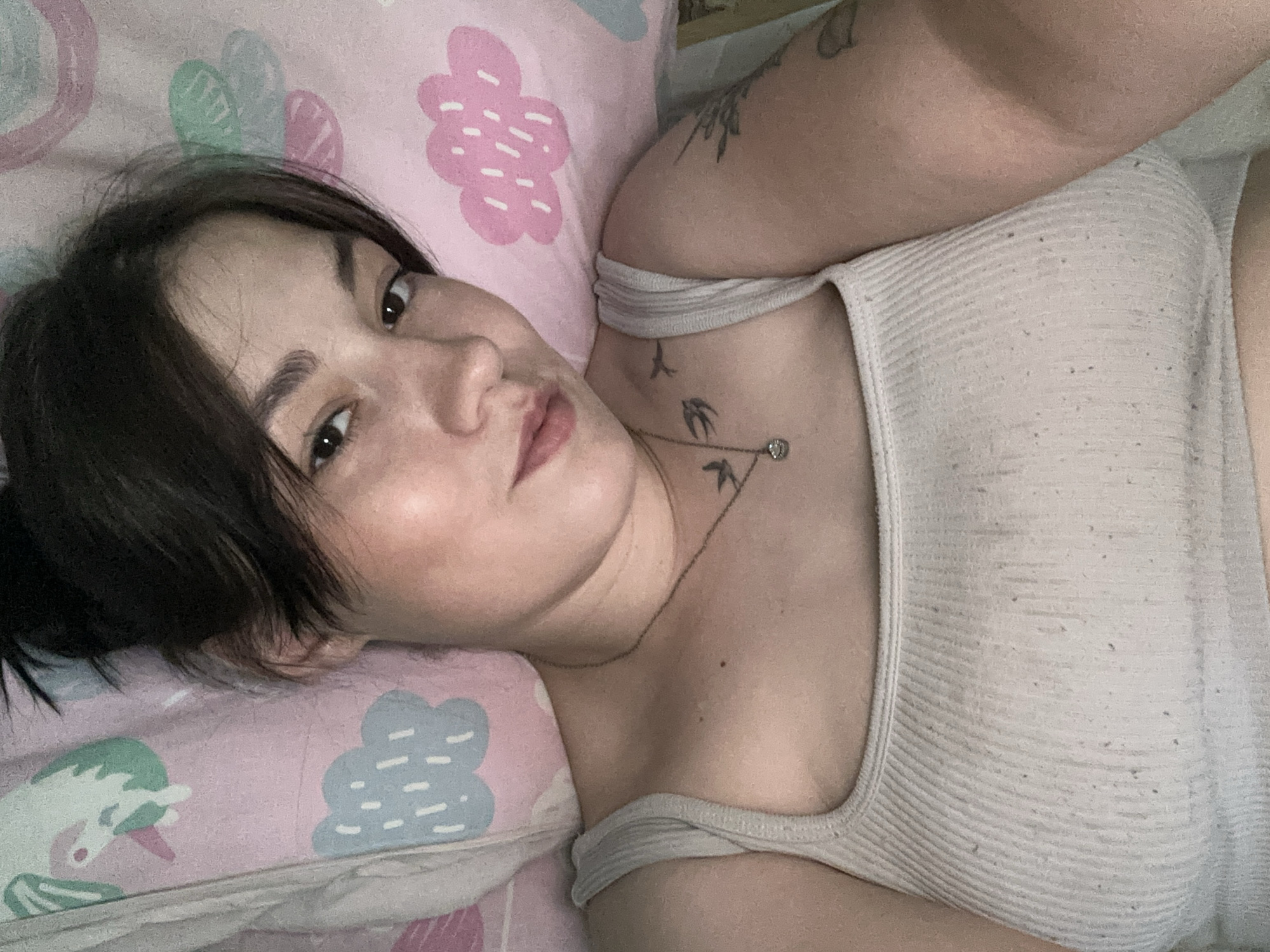 The image shows a selfie of a young woman lying down, possibly in bed, given the pink pillow with cloud and unicorn designs visible. She is wearing a ribbed, cream-colored tank top and a delicate silver necklace with a small pendant. Her dark hair is pulled up, and she has a tattoo on her left arm. The lighting is soft, suggesting an indoor setting. There are no discernible elements in the image that would indicate the specific location within Chaikovsky, Russia.