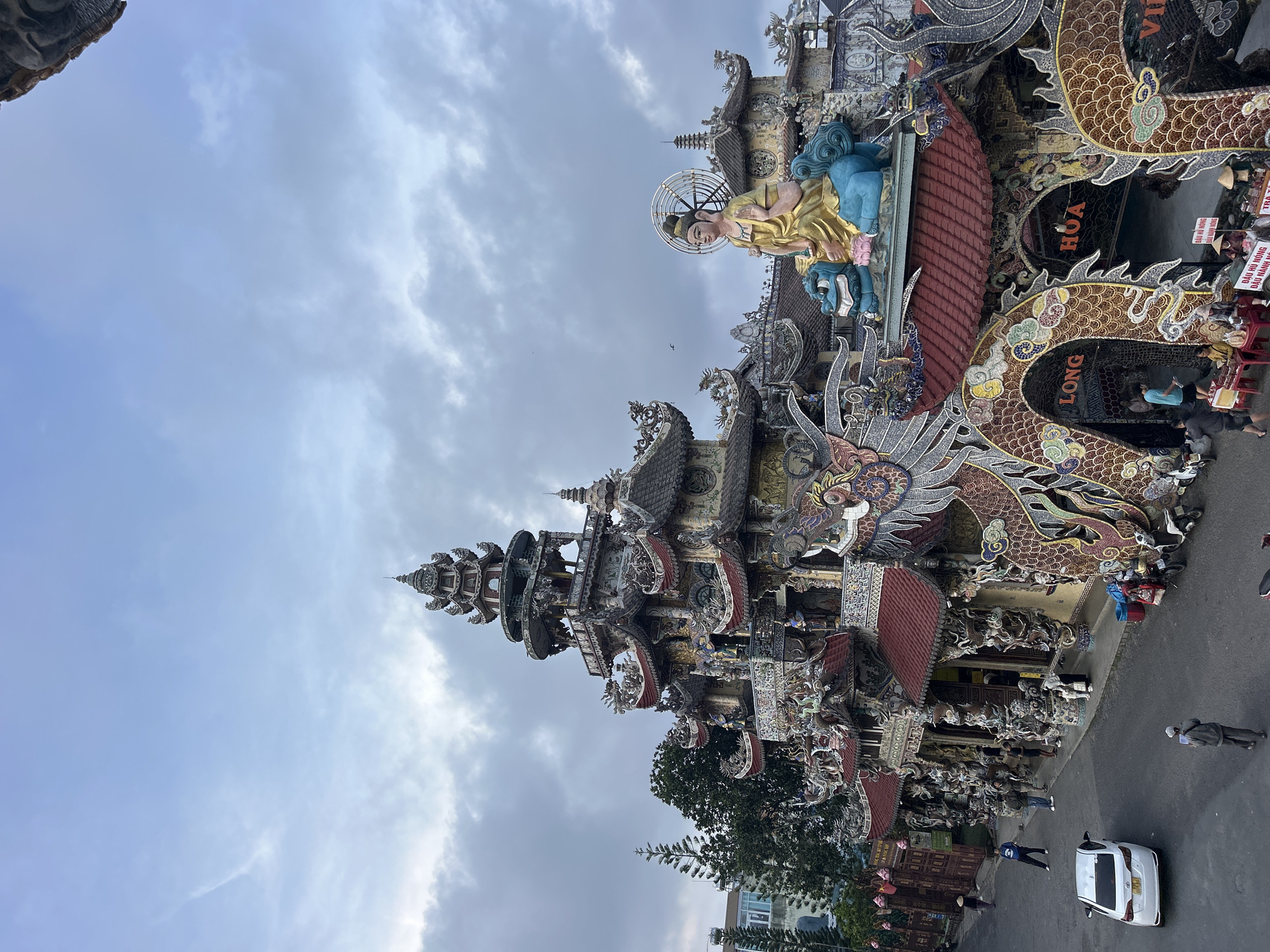 This vibrant image depicts a richly decorated temple or pagoda complex in Xuân Thành, Vietnam, captured under an overcast sky. The central subject is a multi-tiered building, densely adorned with intricate mosaic tiles, sculptures, and reliefs that portray mythical creatures and divine figures.
A striking feature is the large golden statue, possibly of a Buddha or Bodhisattva, seated on a blue base on an upper level, framed by a radiant halo. Below it, elaborate dragon sculptures, crafted with colorful scales and flowing forms, coil around pillars and archways. One prominent dragon sculpture clearly displays the word "LONG," while another section of the building reads "HOA."
On the ground level, several people are visible, some strolling casually, others gathered near what appear to be street vendors or seating areas. One individual wears a traditional Vietnamese conical hat (nón lá). A white car is parked on the street adjacent to the temple grounds.
Visible text on signs includes "ĐẬU HŨ NÓNG" (Hot Tofu) and "DẦU NÀNH NÓNG" (Hot Soy Milk), indicating the presence of food and beverage stalls. Partial text like "VI" and "Co" can also be seen. The scene captures the unique architectural grandeur and cultural activity of a Vietnamese spiritual site during daylight hours.