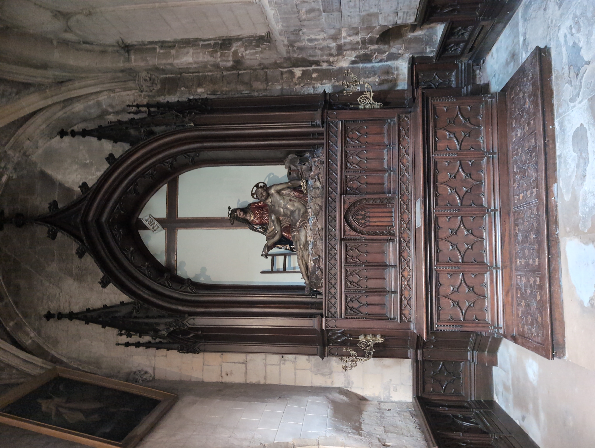 This is a full-shot, eye-level indoor photograph of a religious monument within the church of Saint-Jacques in Le Tréport, France. The monument is ornate and made of dark wood, with intricate Gothic detailing. At the top of the monument, within a niche, are two carved figures. The figure on the right is a male, lying down, appearing to be Christ on the cross, as evidenced by his pose and wounds. The figure on the left is a female, seated, appearing to be the Virgin Mary, cradling the male figure. Behind them, a white cross with the letters "INRI" is visible. Below the niche, a tiered wooden structure features elaborate carvings, including quatrefoil patterns and vertical fluting. The monument is set against a stone wall, and to the left, a framed painting is visible on the wall. Two brass candelabra are mounted on the stone wall on either side of the monument. The floor in front of the monument is tiled with stone and shows some signs of wear. The overall lighting is dim, typical of an interior church setting.