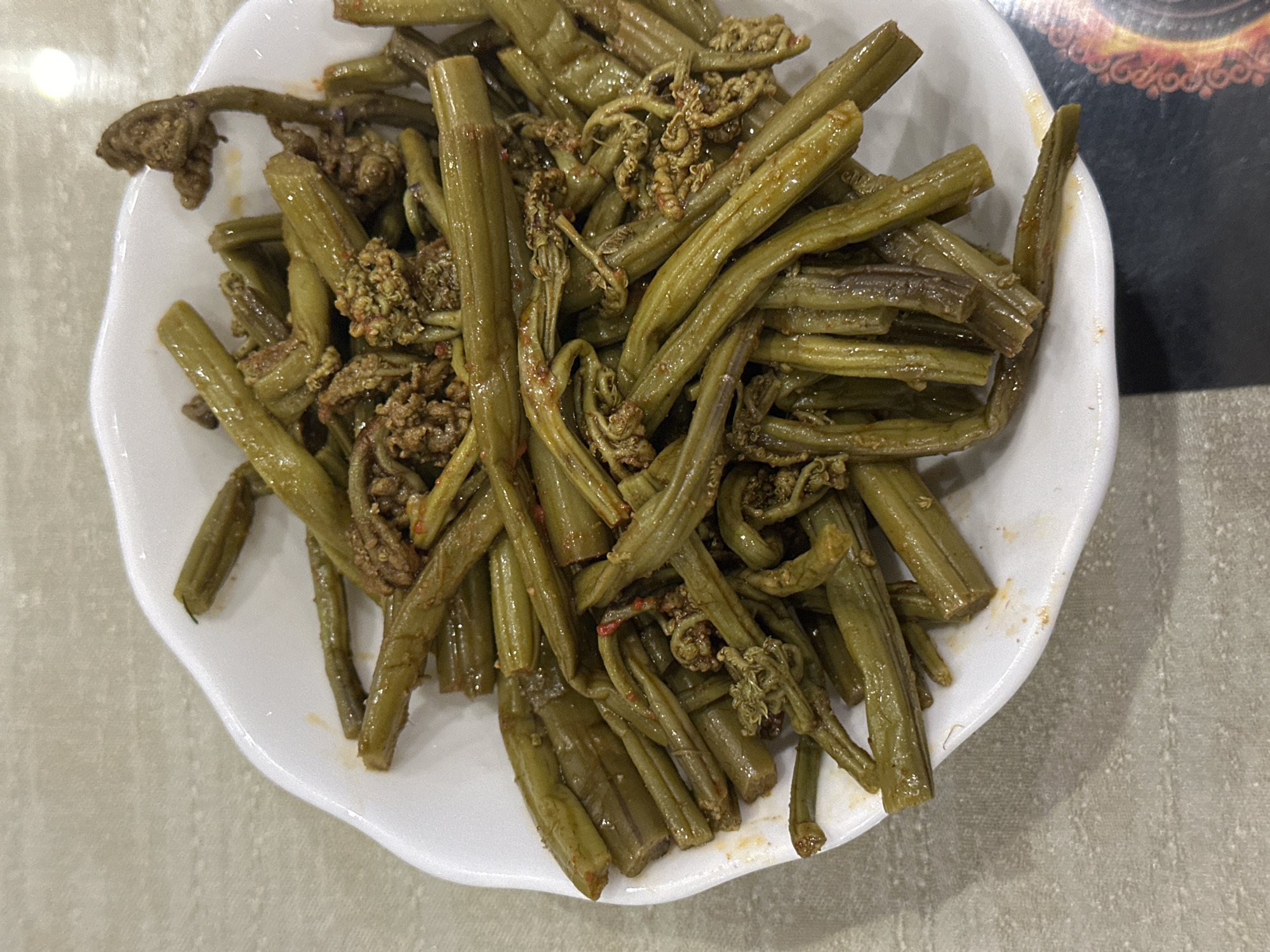 A close-up, high-angle, overhead shot features a white bowl filled with cooked fiddleheads. The fiddleheads are a deep green with some reddish-brown tips, and appear to be coated in a light sauce or oil, with flecks of red seasoning visible throughout. The bowl is slightly tilted, with some of the contents spilling over the edge onto a textured, light-colored tablecloth. The background is out of focus, suggesting the image was taken indoors, possibly in a restaurant. There are no people visible in the image. The scene appears to be set in the city of Nagornyi, Russia.