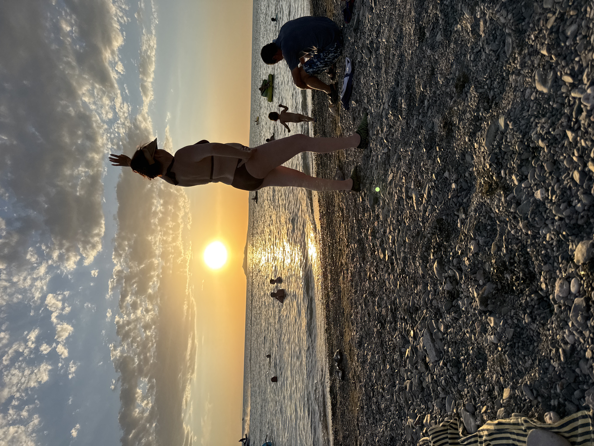 A low-angle, full shot captures a beach scene at sunset. The sun, a bright orange orb, is positioned in the upper left quadrant, casting a warm glow across the sky and water. Wispy clouds dot the sky, tinged with orange and yellow hues. The ocean is calm, with gentle waves lapping at the shore. Several people are visible in the water, swimming and enjoying the evening. In the foreground, a pebbly beach is strewn with rocks and a few items of clothing, including a striped blanket. A woman in a bikini and visor stands facing away from the camera, with her right arm raised. To her right, a man in a blue shirt and patterned shorts sits on the beach. A small child runs towards the water in the middle ground. In the background, a jet ski speeds across the water. The overall impression is one of a relaxed summer evening at the beach. This scene is located on the shore of the Urdzhar reservoir in the city of Urdzhar, Kazakhstan.