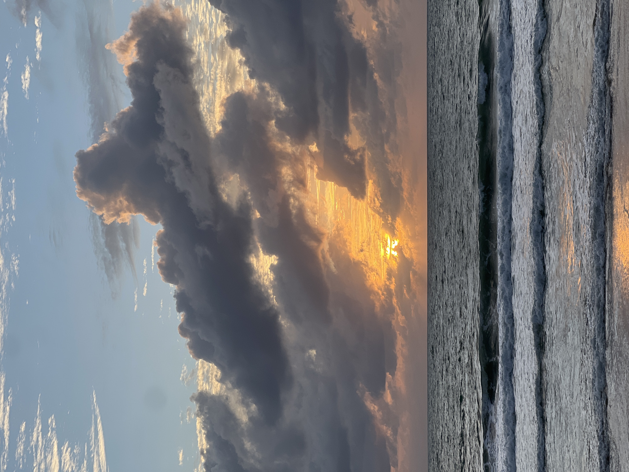 A sunset over the ocean is captured in this image. The sun is partially obscured by dark, dramatic clouds, with rays of light breaking through, creating a warm glow. The ocean in the foreground is dark and choppy, with several waves crashing onto the shore. The sky is a mix of blue and grey clouds, with hints of orange and pink from the setting sun. The scene appears to be a beach in the city of Choeng Thale, Thailand.
