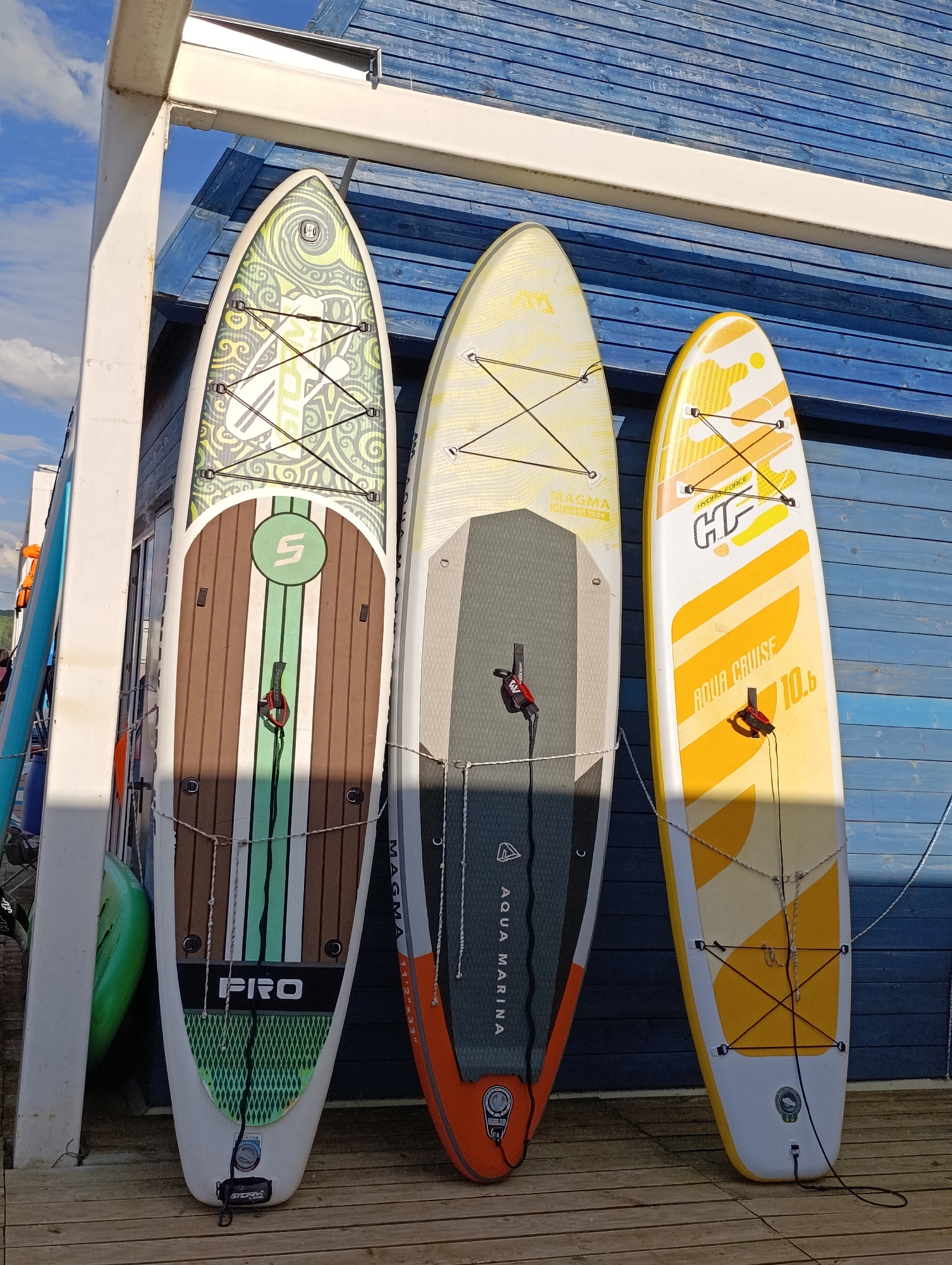 Three stand-up paddleboards are leaning against a blue wooden wall. The board on the left has a green and white tribal pattern at the top, a brown wood-grain pattern in the middle with green and white stripes, and a green diamond pattern at the bottom with the word "PRO" in white letters. The middle board is white with yellow patterns and a gray section at the bottom. The right board is yellow with white patterns and the words "AQUA CRUISE 10.6" printed on it. All boards have bungee cords for securing gear and handles. A green kayak is visible to the left of the paddleboards. The ground is made of wooden planks. The sky is blue with white clouds.