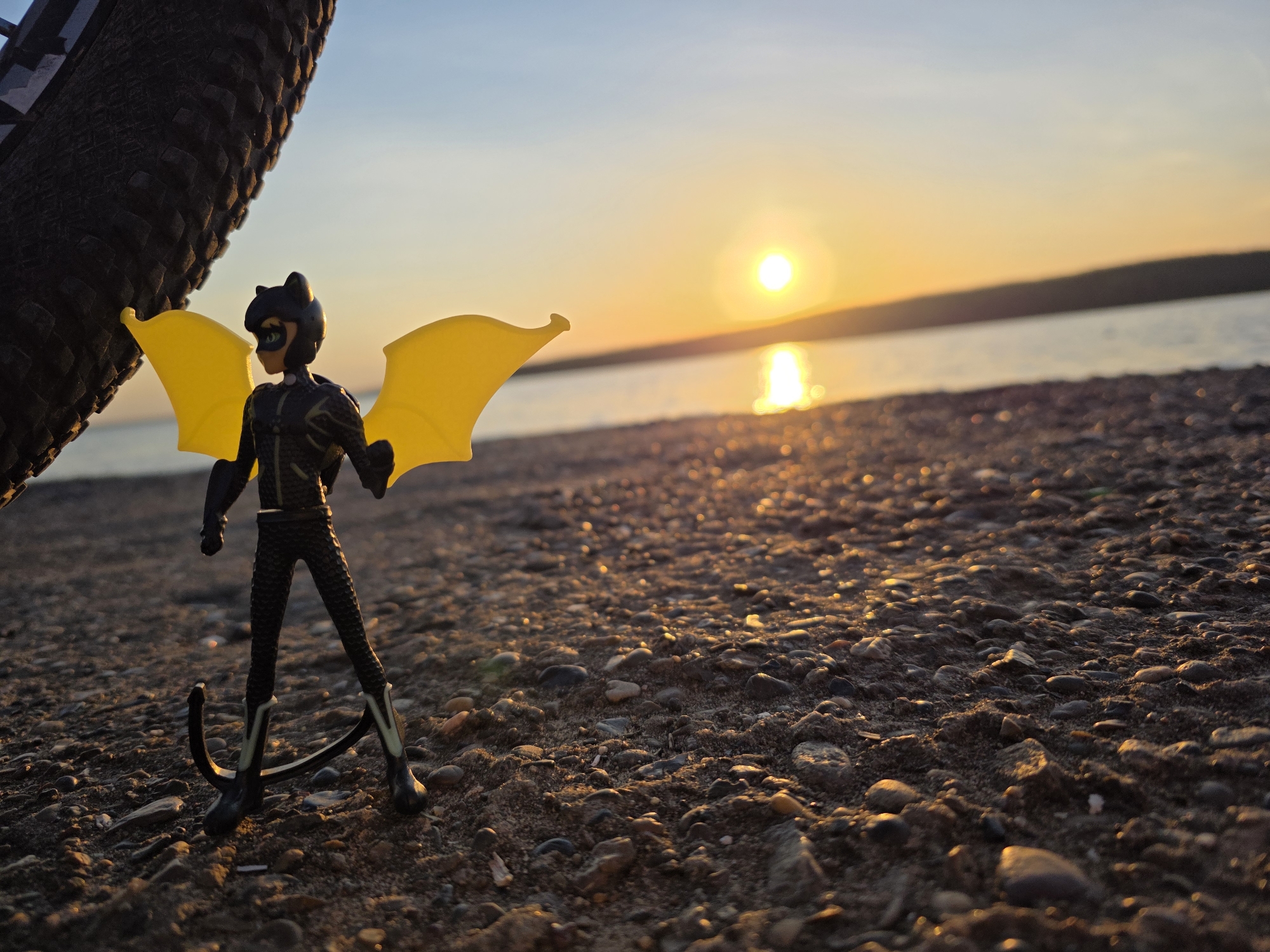 The image shows a toy figure of a character with yellow wings, standing on a pebble-strewn beach next to a bicycle tire. The background features a body of water reflecting the light of the setting sun, with land visible in the distance. The location is likely near Chaikovsky, Russia, based on the request. The figure is primarily black with yellow details and appears to be posed for a photograph. The overall scene evokes a sense of calm and adventure at sunset.
