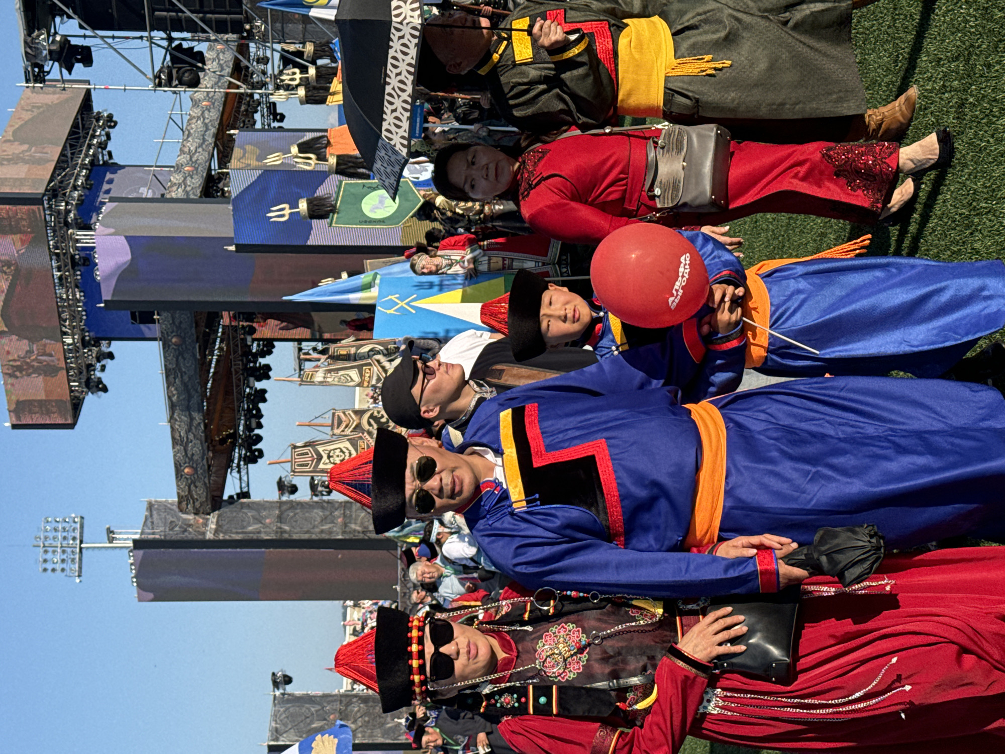 The image depicts a group of people in traditional attire at an outdoor event, possibly a cultural festival or parade. The subjects are dressed in vibrantly colored garments, including robes and headdresses, suggesting a celebration of heritage. In the foreground, several individuals are visible, some wearing sunglasses, indicating a bright, sunny day. One person holds a red balloon. In the background, there are large screens and stage structures, typical of an event venue. The overall atmosphere appears festive and engaging. The location is identified as Ulan-Ude, Russia. The visible text on the red balloon reads "ВЫГОДНО" which translates to "profitable" or "advantageous."