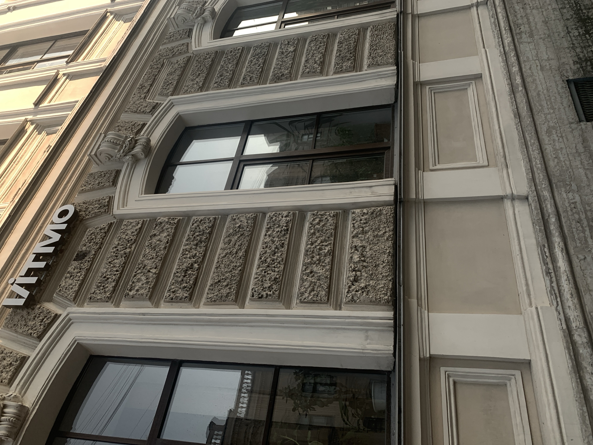The image depicts a section of a building's facade in Saint Petersburg, Russia. The building features architectural elements such as decorated window frames, textured walls, and a vertical sign reading "ИТМО" (ITMO), which stands for Saint Petersburg National Research University of Information Technologies, Mechanics and Optics. The building appears to be multi-story with varying window designs. The environment appears to be an urban setting, suggested by the adjacent buildings and daylight.