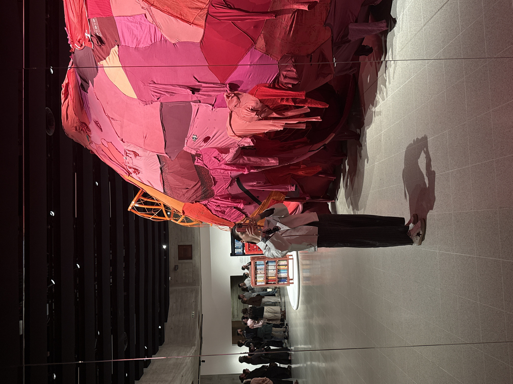 The image depicts the installation "Entwined: Freedom to Move" by Yinka Shonibare CBE RA, located in the Turbine Hall of the Tate Modern in London, United Kingdom. A large, bulbous sculpture composed of red and pink fabrics hangs from the ceiling, partially obscuring the view of the Turbine Hall's structure. Below the sculpture, a crowd of people are observing the installation. A person in a white jacket and dark pants is taking a photograph of the sculpture. A bookcase filled with colorful objects is visible behind the person. The floor is made of square tiles. The ceiling above features a series of dark, vertical panels. The shadow of a person taking a photograph is cast onto the floor.