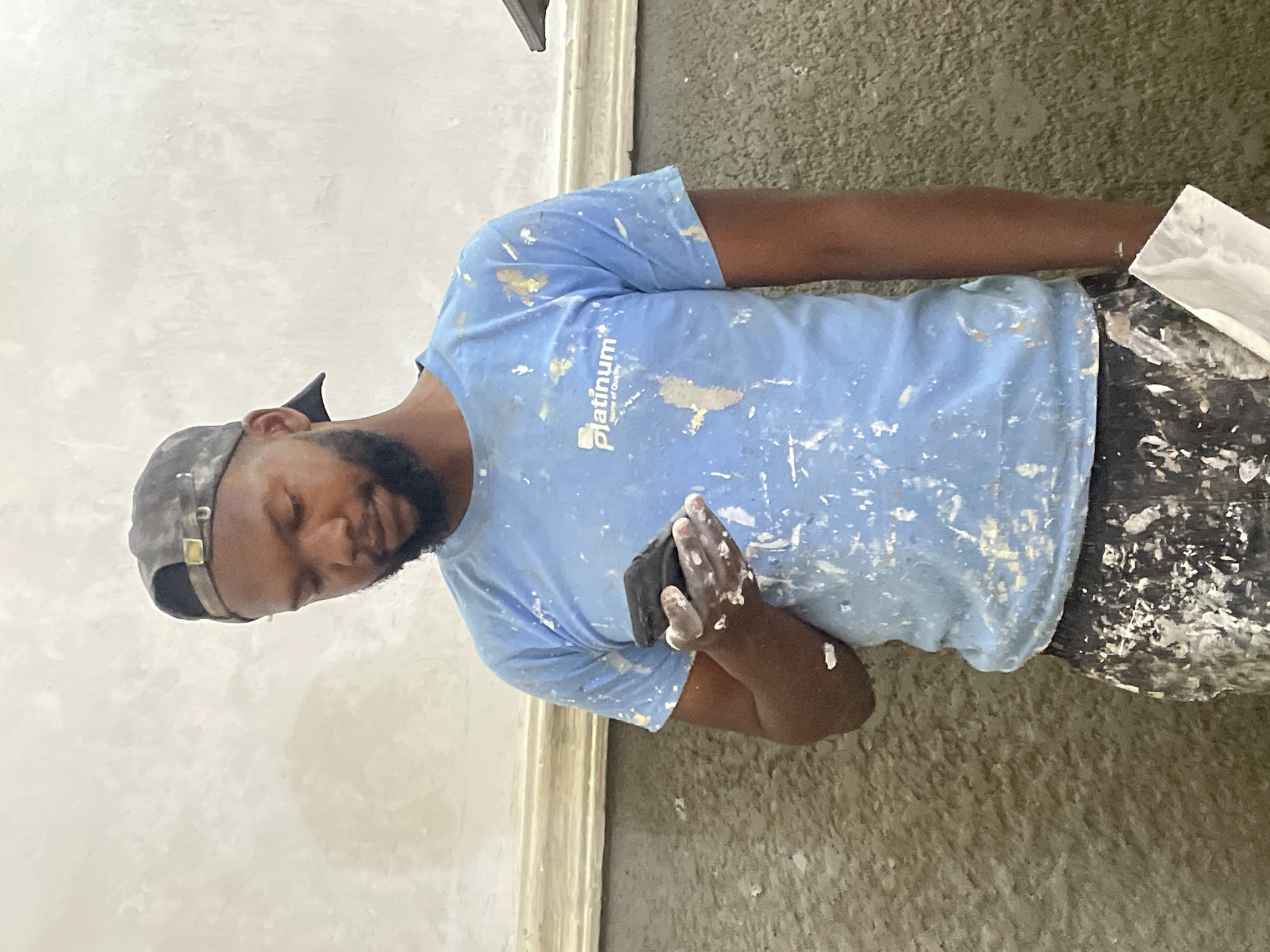 A man wearing a light blue t-shirt and a dark baseball cap is looking down at his phone, which he holds in his right hand. His t-shirt and pants are splattered with white paint. His face is partially obscured by the angle, but he appears to have a beard and is looking intently at the screen. His left hand is also covered in white paint. He is standing in front of a textured, light-colored wall with a decorative molding running horizontally. To his right, a gray, rough-textured wall is visible. The overall environment suggests a construction or renovation site. The lighting is somewhat dim, casting soft shadows.