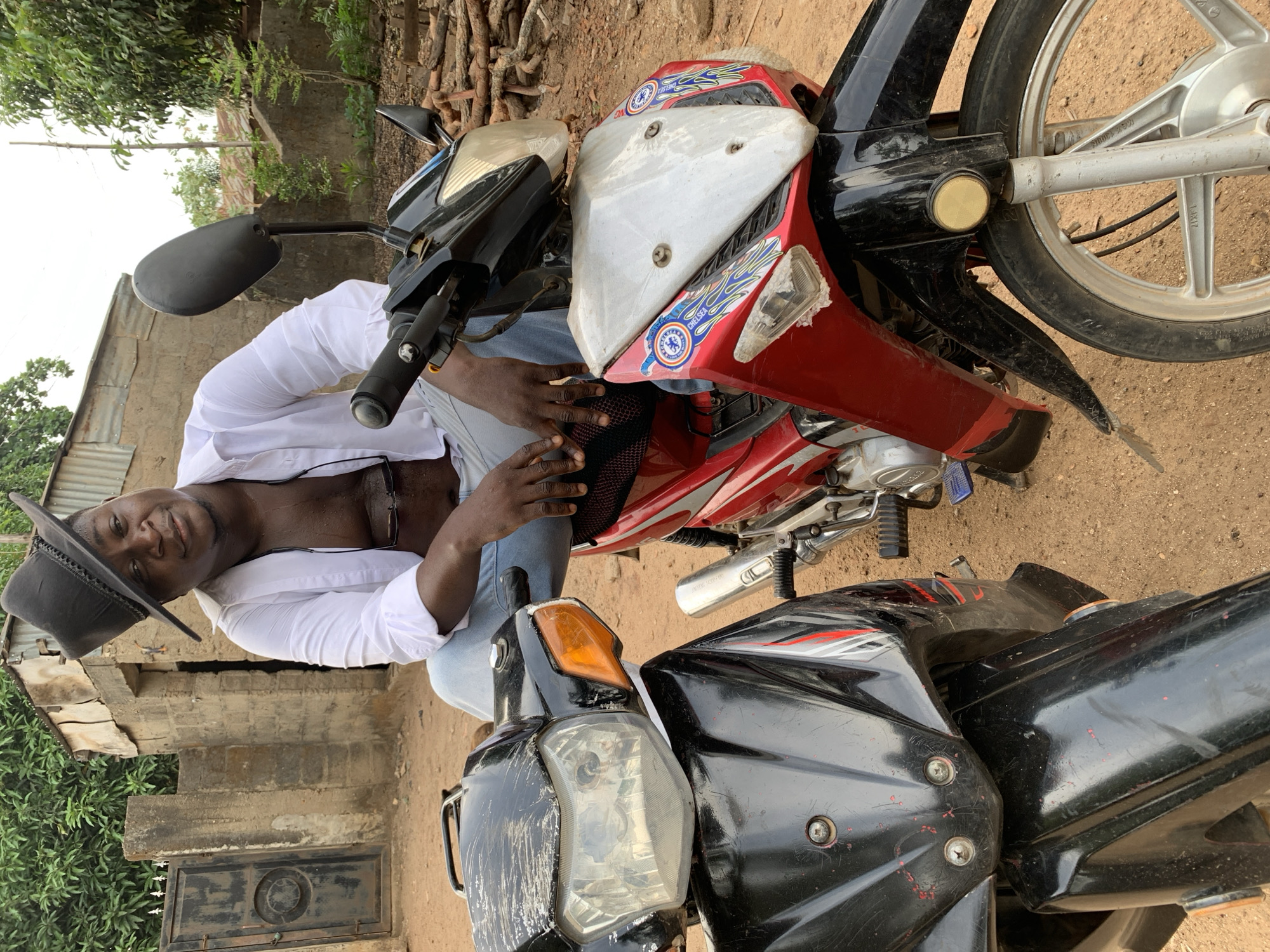 An outdoor, daylight scene in Akwanga, Nigeria, captures a man posing with two motorcycles. The image is rotated counter-clockwise.
The central figure is a dark-skinned man wearing a white long-sleeved shirt, unbuttoned to reveal his chest, light blue jeans, and a dark cowboy hat. He has glasses hanging from his shirt collar and a slight beard. He is seated on a red motorcycle, resting his hands casually on its black seat, and looks towards the camera with a neutral to slight smile.
The motorcycle he is on is predominantly red with white and black accents. Its fairings are decorated with "Chelsea" football club stickers and blue flame decals. A silver exhaust pipe and a silver spoked wheel are visible, with "MAX 11000PM" and "1XM17" inscribed on the wheel.
Partially in the foreground to the man's right is another motorcycle, mostly black, with red accents. This bike appears older and shows numerous scratches. Its front headlight, an amber turn signal, a black rearview mirror, and handlebars are clearly visible.
The background consists of unpaved dirt ground, green foliage, and rustic concrete block buildings, some with corrugated iron roofs. Piles of logs are stacked behind the red motorcycle. The lighting suggests an overcast sky or soft daylight, as there are no harsh shadows.