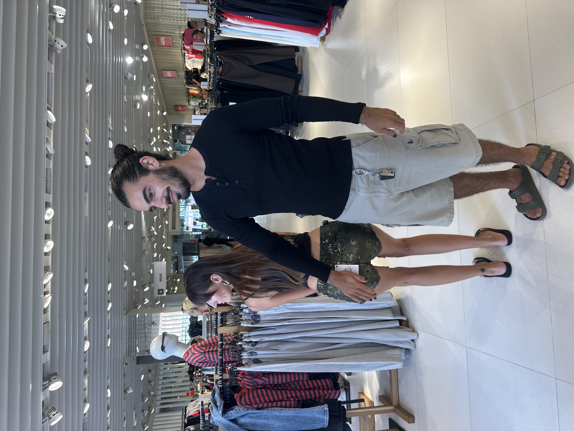 In Kuta, Indonesia, a man with dark hair styled in a bun and a beard stands in a retail store, his right hand on the posterior of a woman. The woman, with long dark hair and hoop earrings, wears a black tank top and gold sequined shorts. She is facing away from the camera, looking to her left. Both individuals are dressed casually. The man is wearing a black long-sleeved shirt and light-colored cargo shorts, accessorized with a keychain hanging from his shorts pocket and dark sandals. The woman wears black sandals and has a tattoo on her left calf.
The background reveals a brightly lit clothing store with racks of garments and mannequins. Signs indicating "MAN" are visible in the distance, along with sale signs displaying "-40%". The floor is tiled in a light color. The overall environment suggests a shopping center or department store.