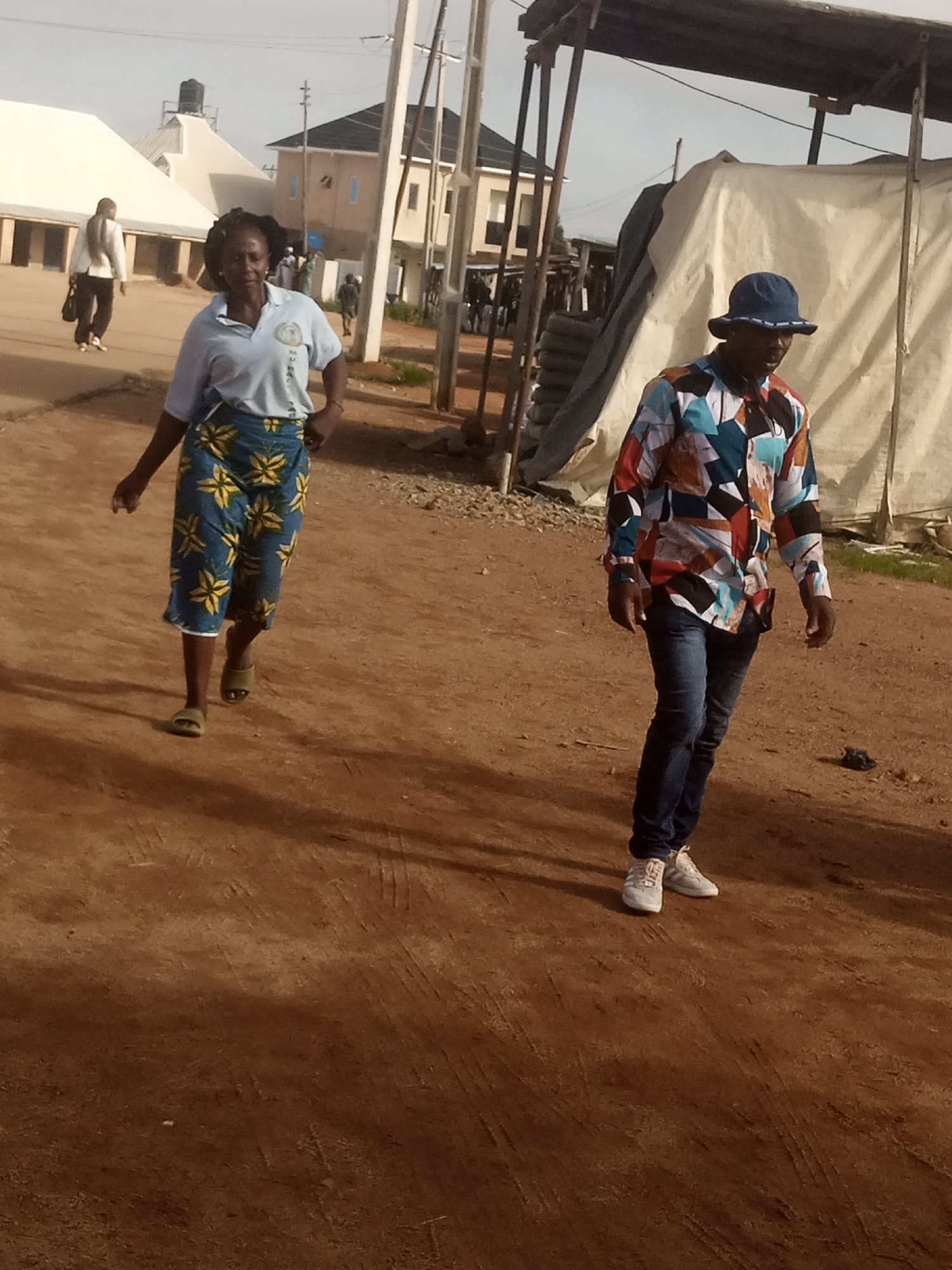 In Katon Rikkos, Nigeria, a man in a colorful, patterned shirt and blue jeans walks on a dirt path. He wears a blue bucket hat and white sneakers. To his left, a woman in a light blue polo shirt and a blue skirt with a yellow floral pattern walks in the opposite direction. She is wearing open-toed sandals. Behind them, a white tent structure and buildings are visible, suggesting a marketplace or gathering area. The ground is uneven and dusty, with tire tracks visible. The lighting indicates it is daytime.