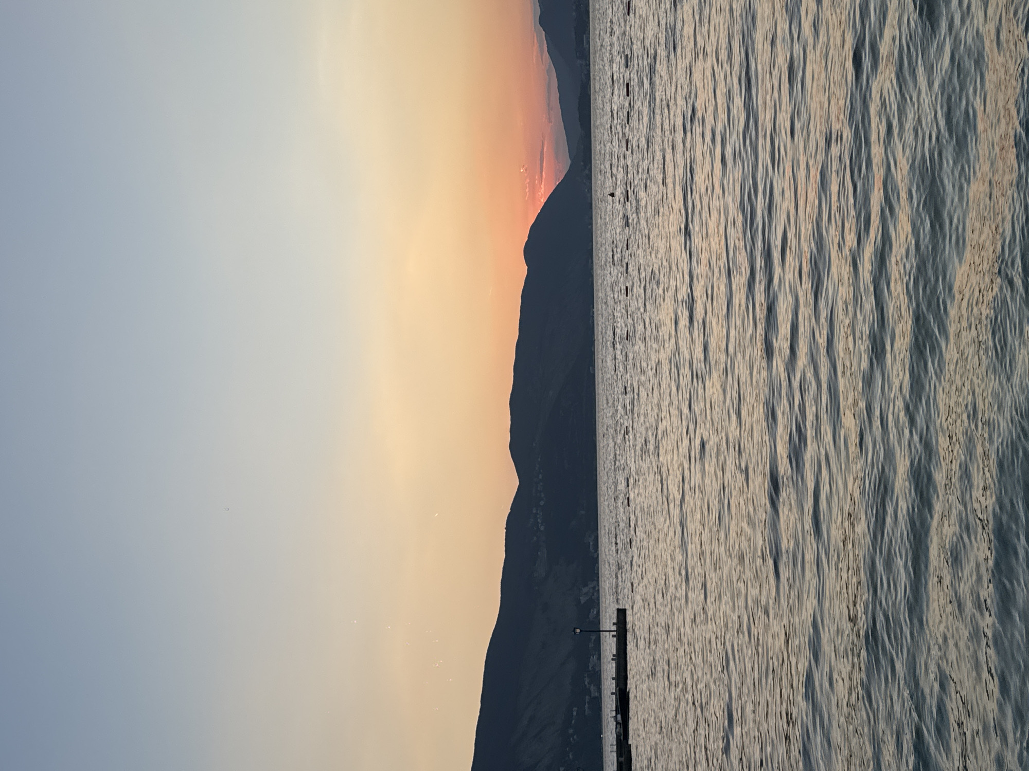 The image depicts a serene seascape at dusk. The sky transitions from a pale blue at the top to a warm orange and pink gradient near the horizon, indicating the sun is setting. Silhouetted against the vibrant sky are dark, undulating mountains. The water in the foreground is calm, with gentle ripples reflecting the colors of the sky. A small pier with a single lamppost extends into the water on the left side of the frame. In the distance, a series of buoys mark a boundary in the water, and a lone figure is visible standing on one of them. The overall atmosphere is peaceful and tranquil.