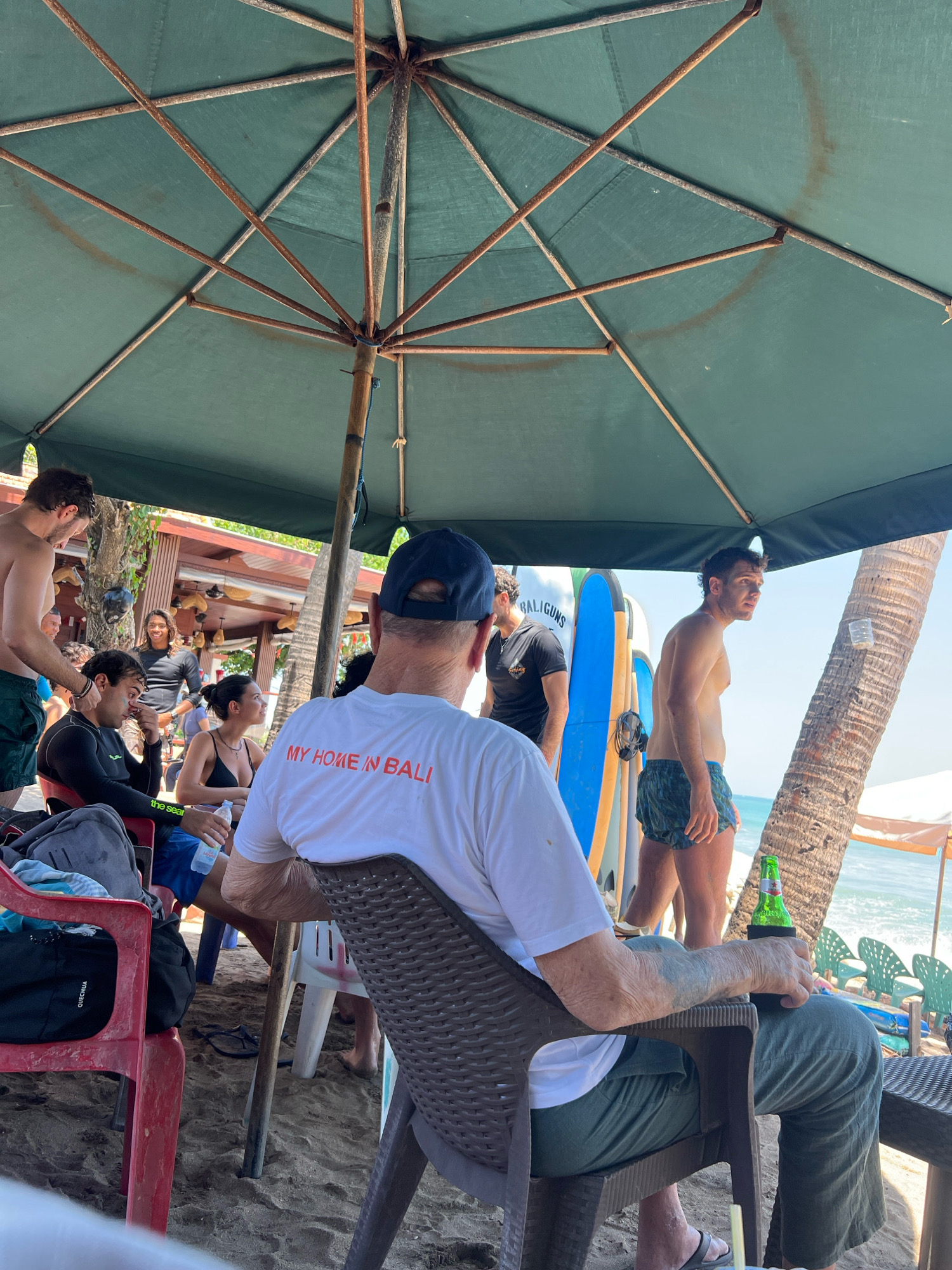 A group of people are gathered on a sandy beach, beneath a large, green, weathered umbrella. The umbrella has a metal frame that appears to be rusting. In the foreground, a man with tattoos on his arm is seated, wearing a white t-shirt with the words "MY HOME IN BALI" printed on the back in red. He is holding a green beer bottle in a holder. To his left, a woman in a black bikini is sitting and holding a bottle of water. Beside her, a man in a black wetsuit is seated. Behind them, several surfboards are propped up against a structure. Another man, shirtless and wearing green patterned swim trunks, stands to the right, looking off into the distance. In the background, the ocean is visible with a clear blue sky overhead. The scene appears to be a casual gathering at a beach bar or restaurant in Kuta, Indonesia.