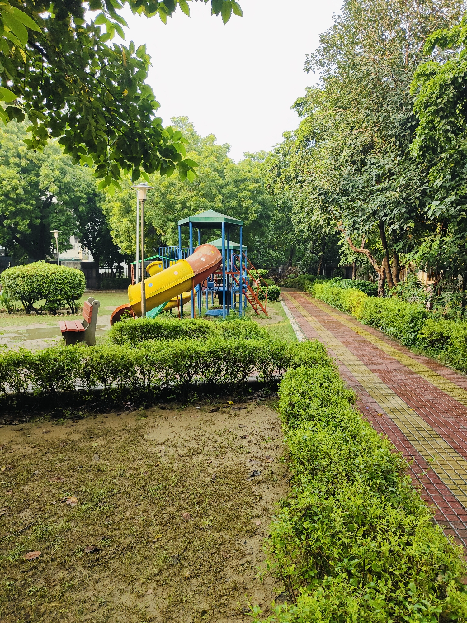 The image depicts a park in Gurgaon, India, featuring a children's playground. The playground equipment includes a bright yellow and red slide, a smaller blue slide, and a climbing structure with red stairs. Adjacent to the playground is a park bench, which appears to be made of wood and has a metal frame. The park features a paved pathway with red and yellow tiles that winds through lush green bushes and manicured hedges. The ground is a mix of grass and damp soil, suggesting recent rain. Tall trees with dense green foliage surround the area, creating a shaded, natural ambiance. The sky is overcast, indicating a cloudy day. There are no people visible in the scene.