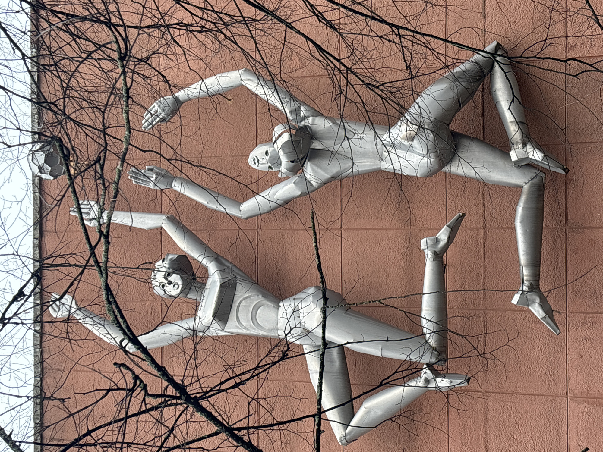 The image shows a public art installation on a building facade. The installation features two stylized, silver-colored figures in dynamic poses, seemingly interacting with a soccer ball. The figures are mounted on a textured, reddish-brown wall that appears to be tiled. Overhanging bare branches obscure parts of the sculpture and the wall. The sculpture is located in the city of Chaikovsky, Russia.