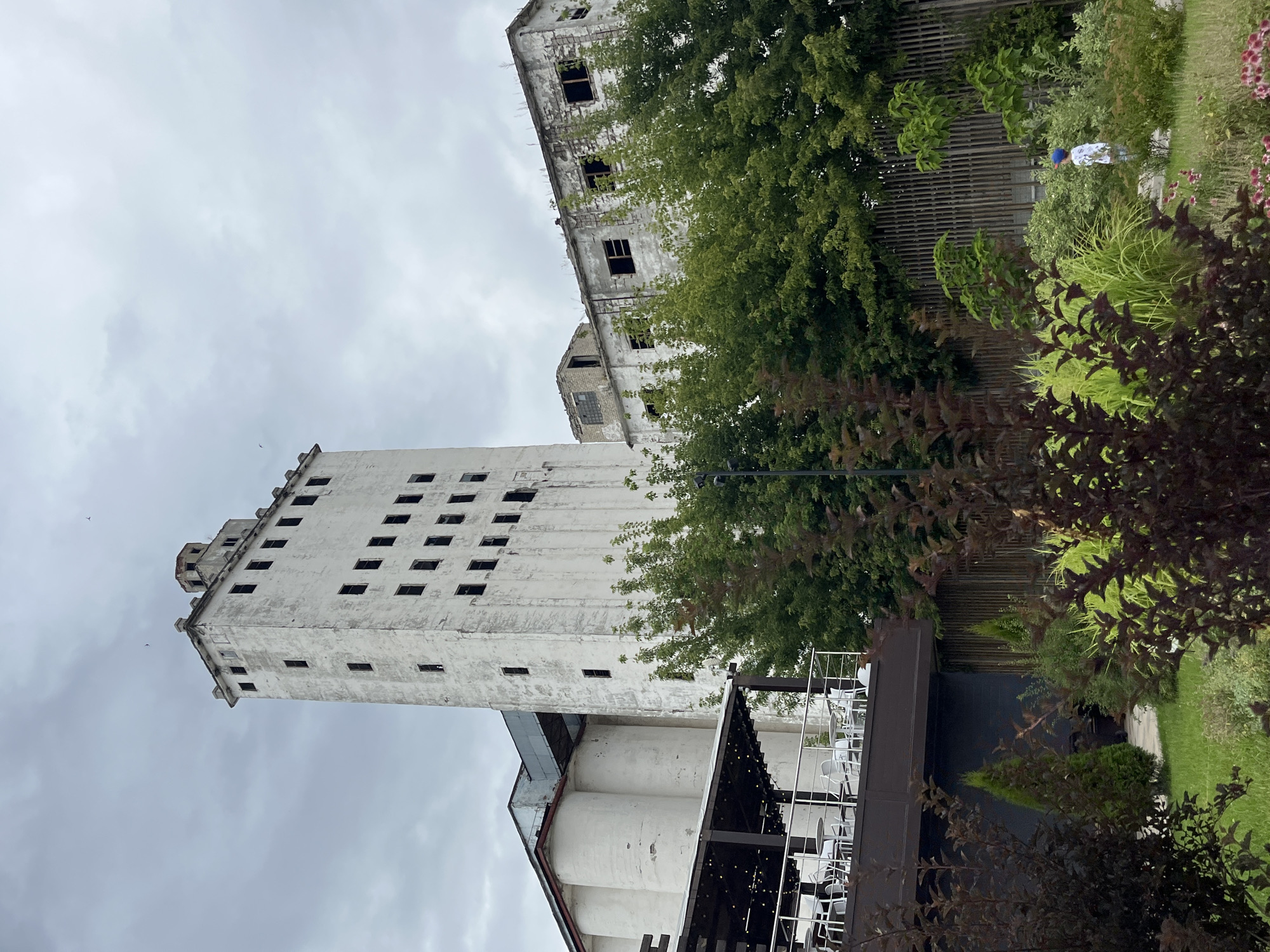 This is a full-frame, eye-level, outdoor, daytime, medium shot of the former Saratov Bread Factory complex, located in Saratov, Russia. The complex consists of a tall, white, multi-story building with rows of dark, rectangular windows and a lower, white, multi-story building with similar window openings. Adjacent to these structures are rounded, white silo-like buildings. In the foreground, a dark-colored elevated deck with tables and chairs is visible, featuring string lights overhead. A small, a child wearing a blue hat and a white shirt is standing in a garden area with various plants and a wooden fence behind them. The sky is overcast with clouds.