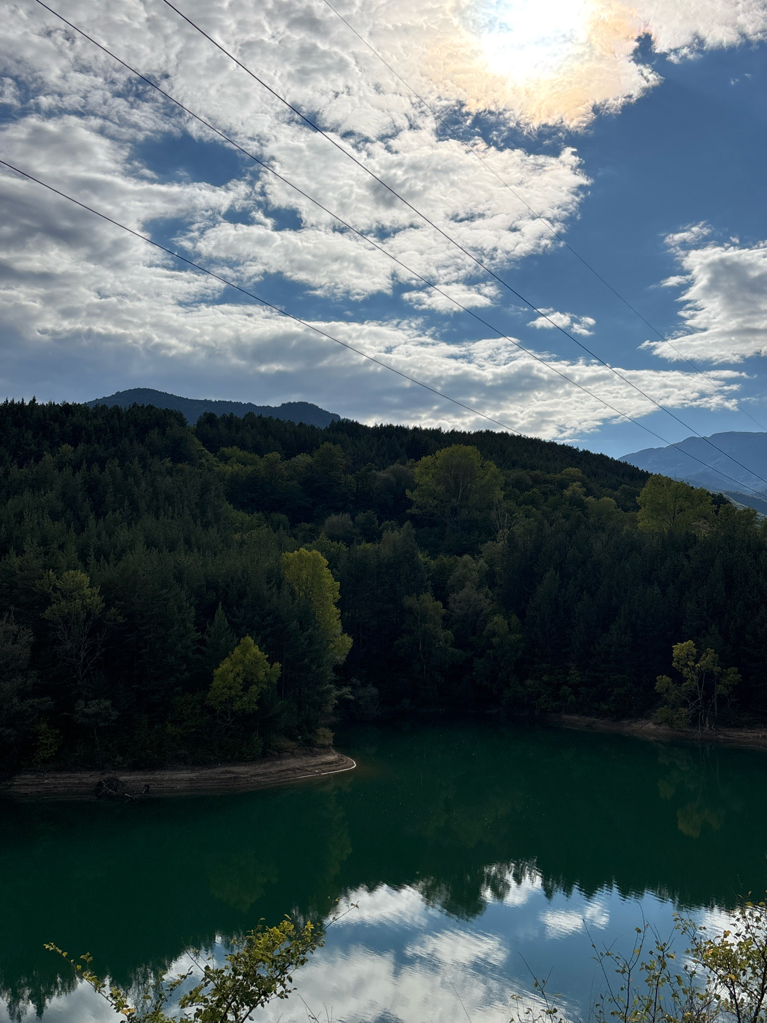 The image captures a serene natural landscape. In the foreground, a body of dark green water reflects the sky and surrounding trees. The water's surface is mostly calm, with subtle ripples. Along the edge of the water, there is a strip of brown earth, and beyond that, a dense forest covers a sloping hillside. The trees are predominantly dark green, with some interspersed lighter green and yellow foliage, suggesting a mix of evergreen and deciduous trees, possibly indicating autumn. In the background, hazy mountain ranges are visible. Above the treeline, a bright sky is filled with a mix of white and gray clouds. Sunlight shines through a gap in the clouds, casting a bright glow in the upper right portion of the image. Several power lines are visible, stretching diagonally across the sky. This scene is located in the vicinity of Dobrotino, Bulgaria.