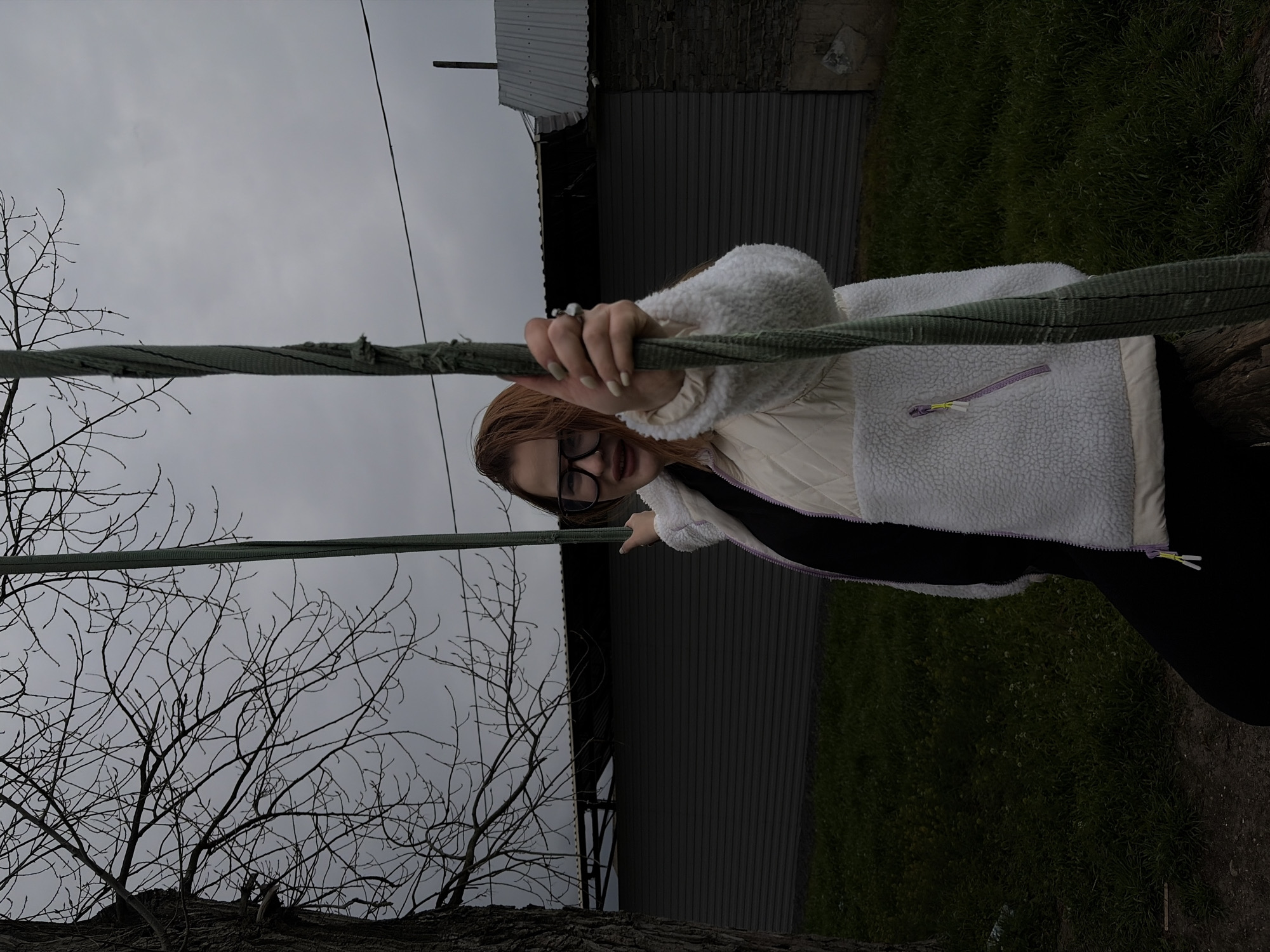 The image displays an outdoor scene in Port-Katon, Russia, under an overcast sky. The primary subject is a young individual, likely female, captured from approximately the chest up. She has reddish-brown hair, wears black-framed glasses, and a nose ring. Her fingernails are manicured with a light polish. She is dressed in a cream or white, textured sherpa-style jacket, layered over a darker garment, with a subtle purple zipper pull visible on a pocket.
Her right hand firmly grasps a thick, worn, green rope-like object that spans horizontally across the mid-ground of the frame. Her left arm is slightly raised, with her index finger pointing towards the left side of the image. Her expression appears neutral, possibly conveying curiosity or engagement.
In the foreground to the left, several bare tree branches, possibly with early buds, suggest a cooler season like late winter or early spring. The background features a large, dark building with a corrugated metal roof and dark siding, possibly a barn or shed. The sky above is a uniform grey, indicative of cloudy weather. To the right, green grass is visible, and a thin vertical pole or line rises into the distance. The overall scene suggests a rustic or semi-rural setting. There is no visible text in the image.